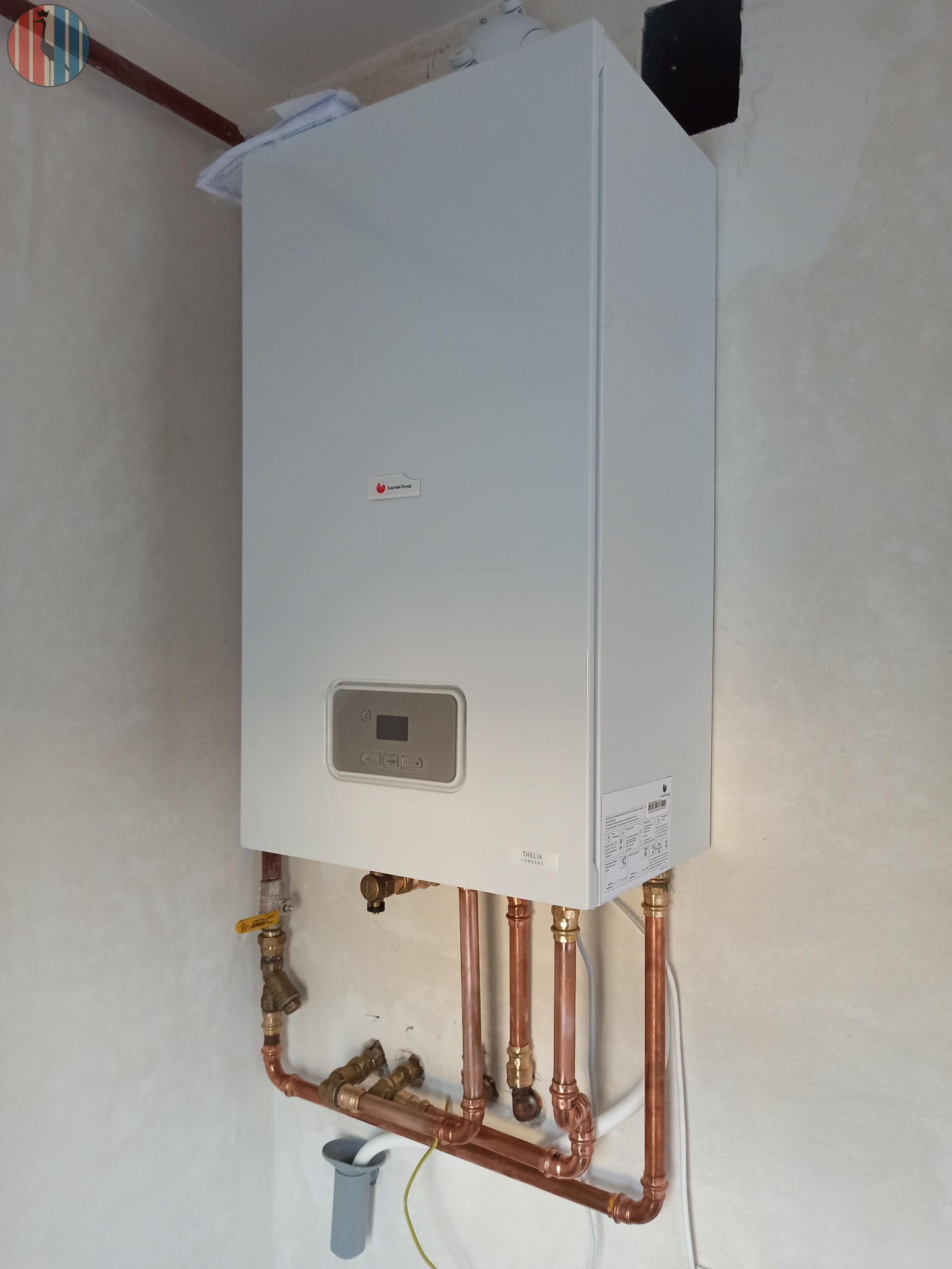Nowy, biały piec gazowy Saunier Duval Thelia Condens zamontowany na ścianie, z widocznymi miedzianymi rurami instalacji, zaworami i szarą rurką odpływową.