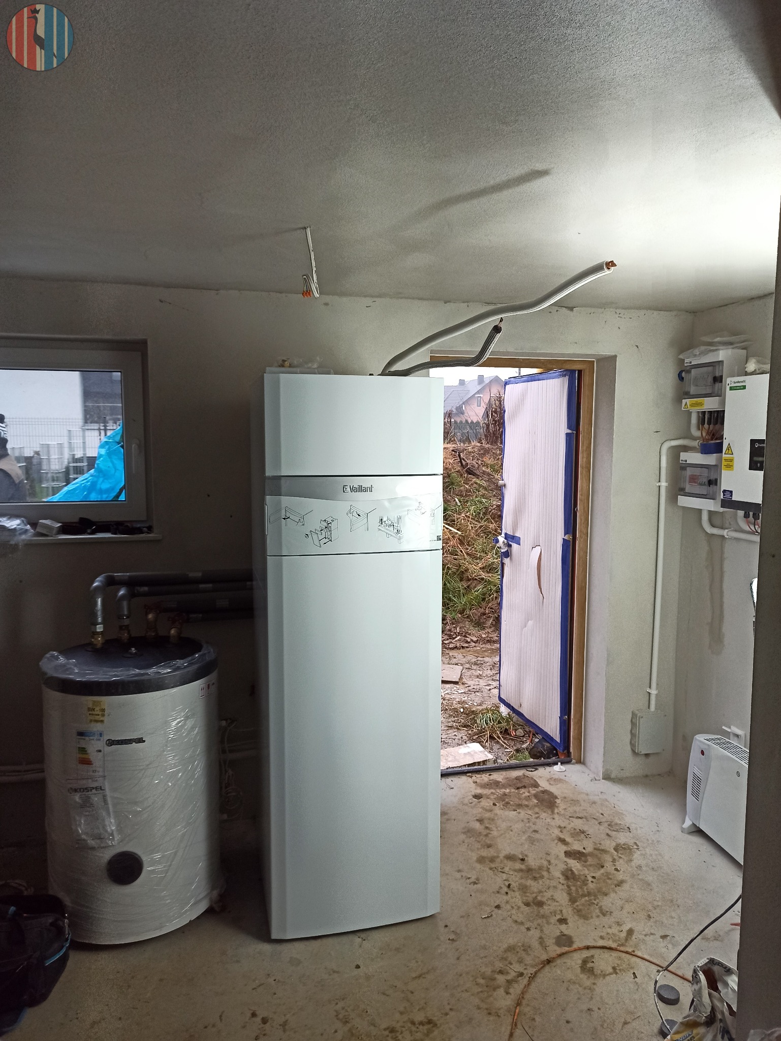 Biały piec gazowy Vaillant i bojler Kospel w folii ochronnej, w surowym pomieszczeniu z betonową podłogą i otwartymi drzwiami na zewnątrz.