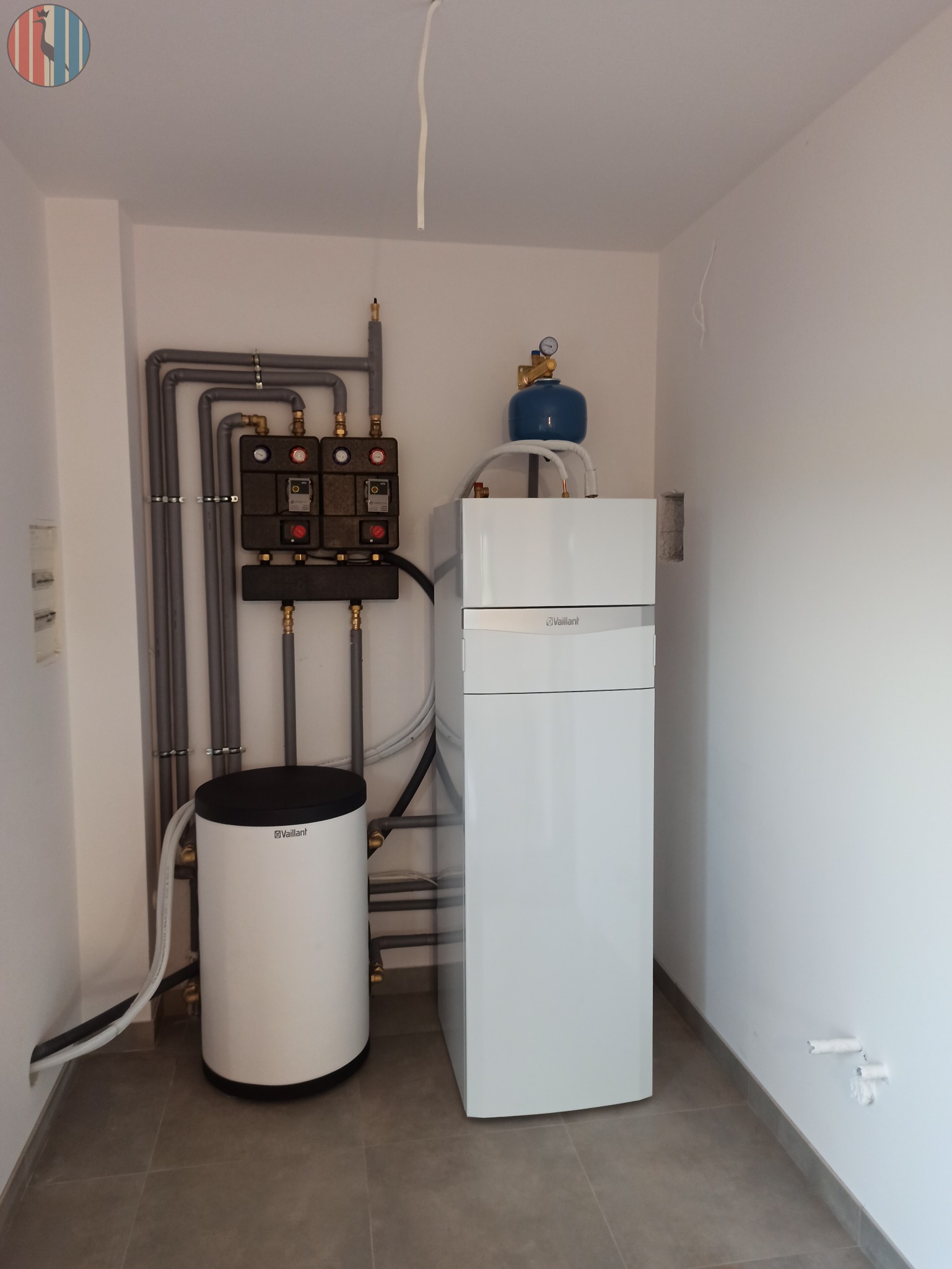 Biały, kondensacyjny piec gazowy Vaillant z niebieskim zbiornikiem wyrównawczym, obok biały zasobnik ciepłej wody, szare rury instalacji grzewczej i dwa czarne rozdzielacze w kotłowni z szarą...