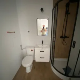 Wykończona łazienka z kabiną prysznicową z czarnymi profilami, białą umywalką z szafką, toaletą i drewnopodobną podłogą; widoczne odbicie osoby robiącej zdjęcie w lustrze.