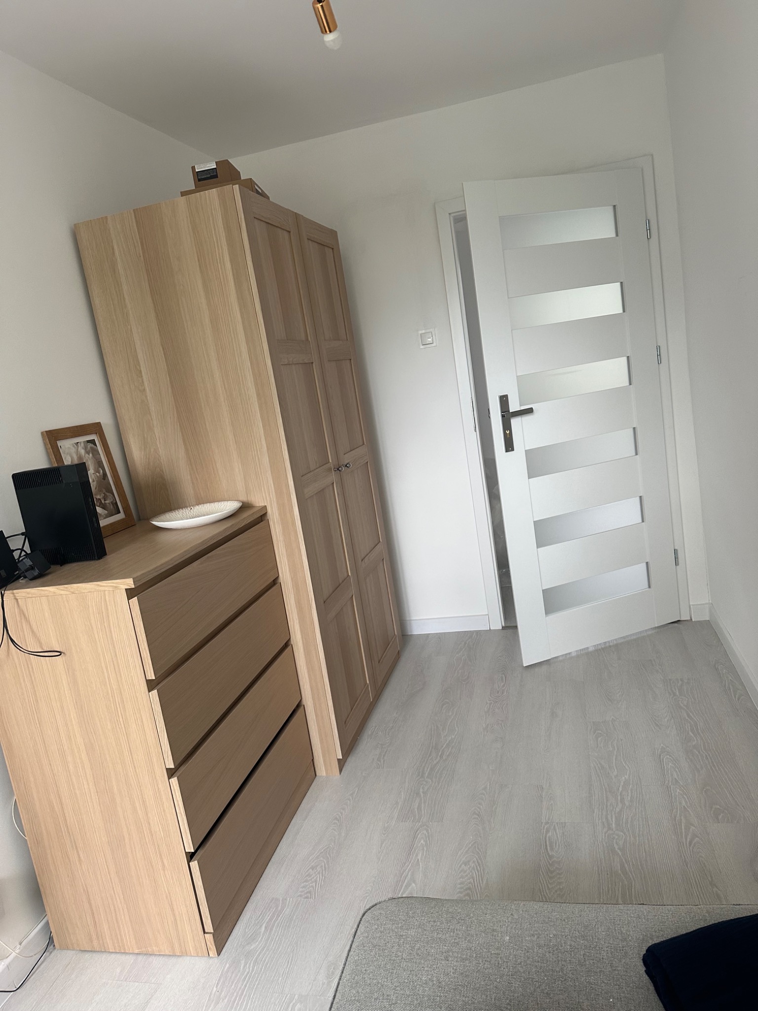 Jasne drewniane meble (komoda z szufladami i szafa) w nowym, minimalistycznym wnętrzu z białymi ścianami, szarą podłogą imitującą drewno i białymi drzwiami z przeszkleniami.