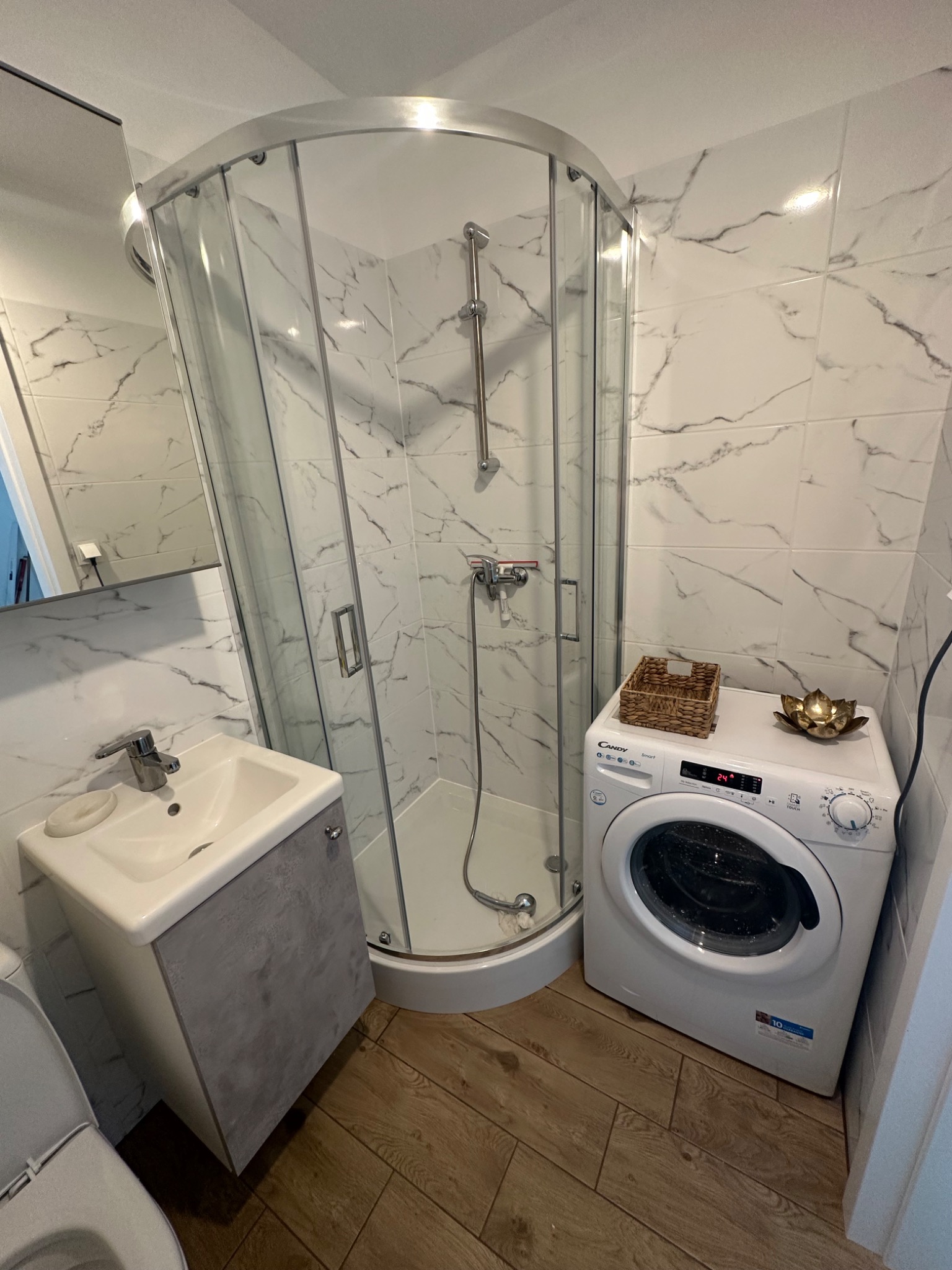 Jasna łazienka z kabiną prysznicową, umywalką z szafką w odcieniach szarości, białą pralką Candy z dekoracyjnym koszykiem i złotą ozdobą na górze, wykończona płytkami imitującymi marmur...