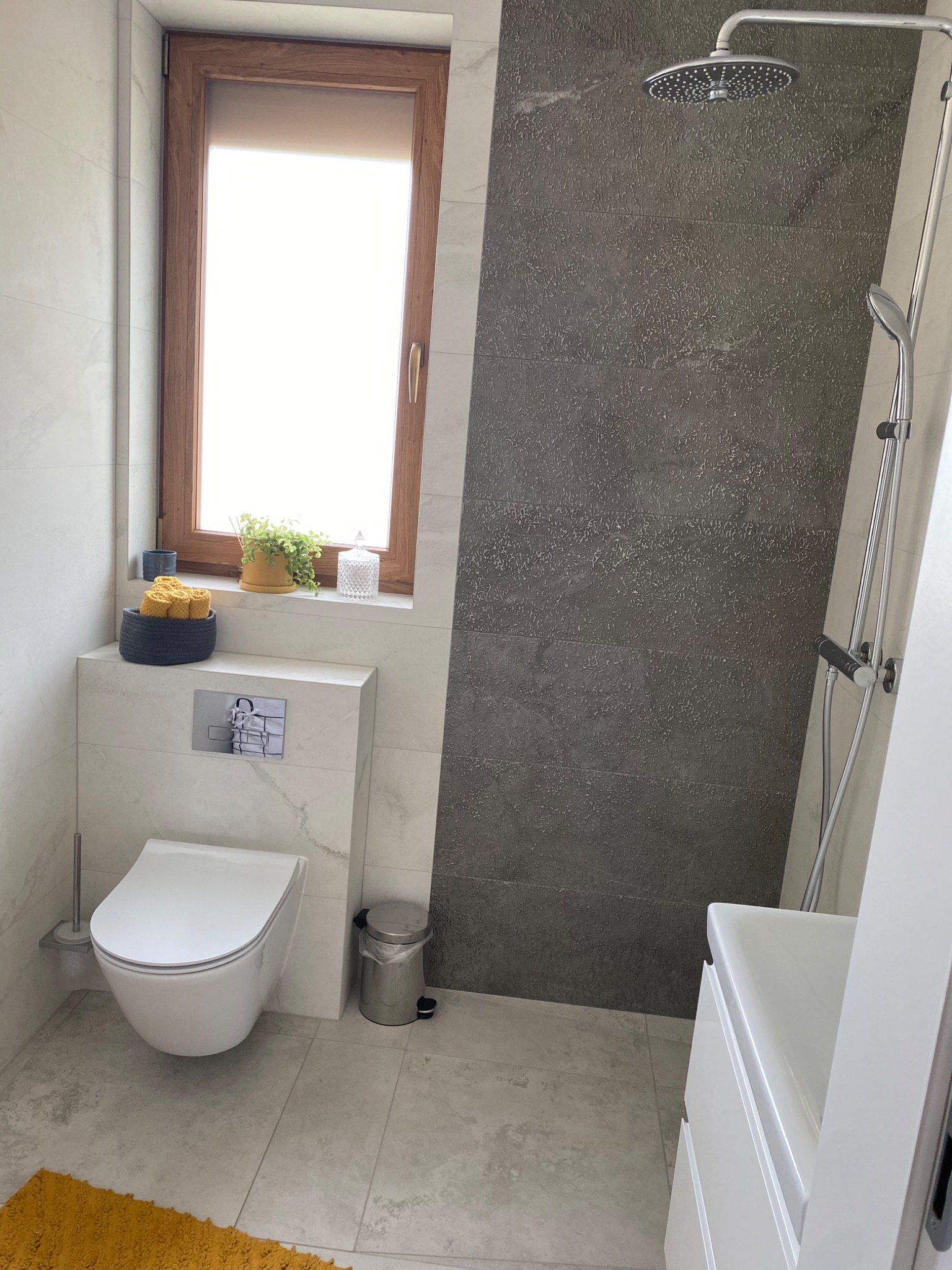 Nowoczesna łazienka z wiszącą toaletą, deszczownicą i oknem nad zabudową geberit, wykończona płytkami w odcieniach szarości i bieli, z akcentami żółtego koloru.