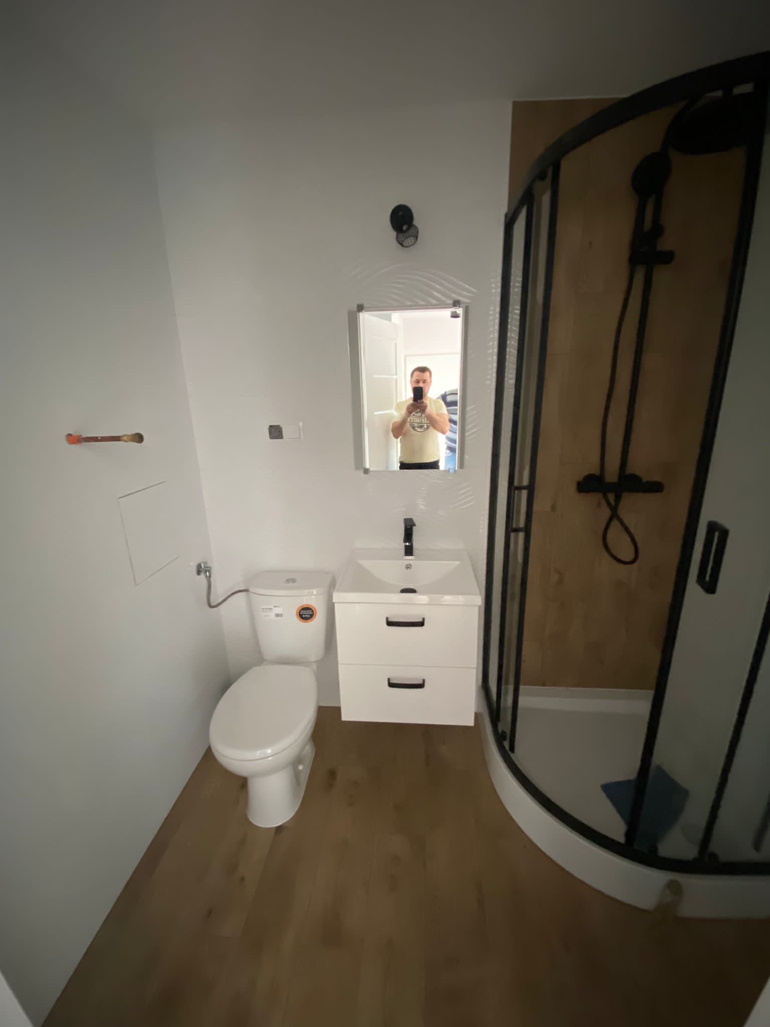 Wykończona łazienka z kabiną prysznicową z czarnymi profilami, białą umywalką z szafką, toaletą i drewnopodobną podłogą; widoczne odbicie osoby robiącej zdjęcie w lustrze.