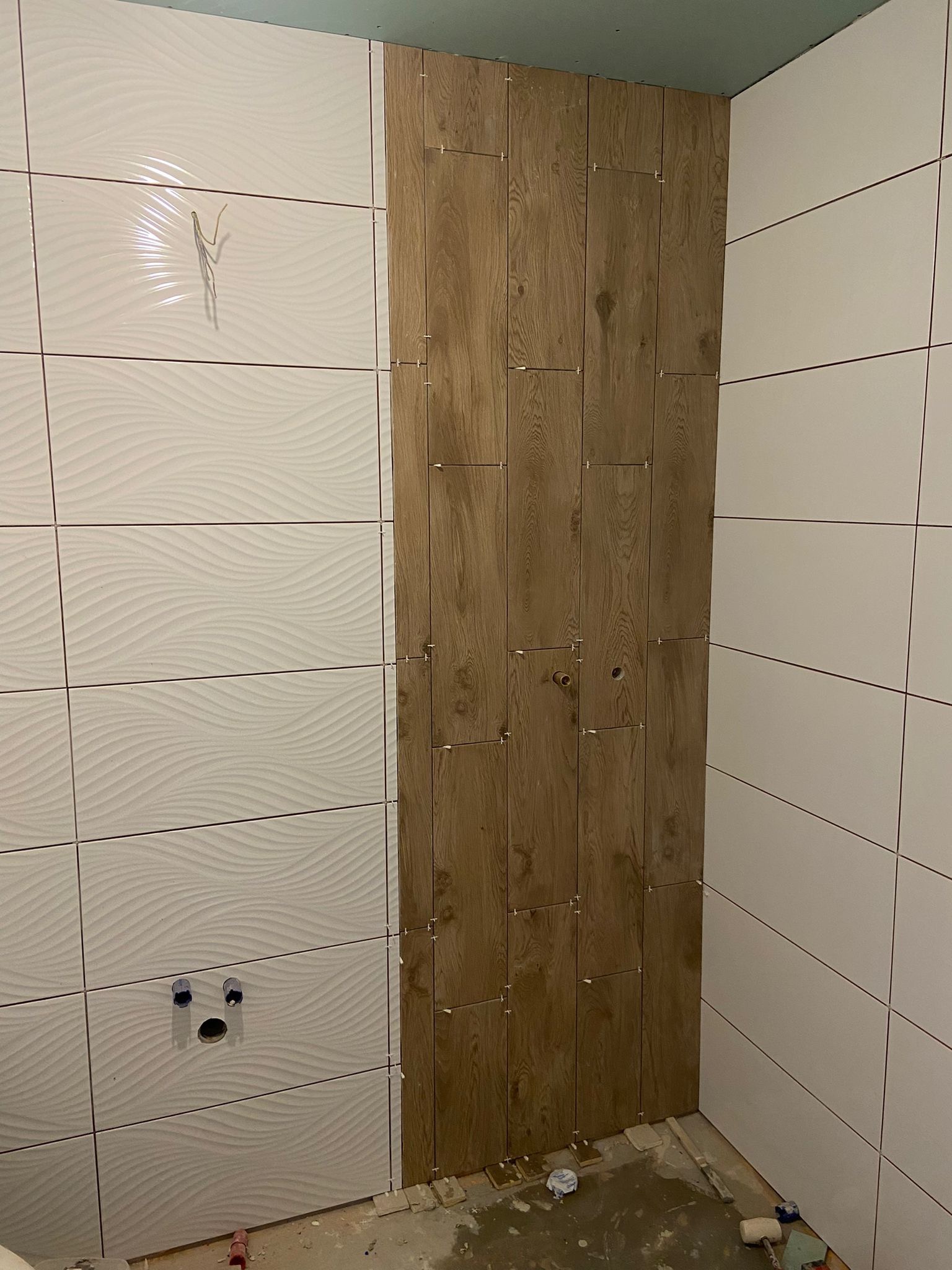 Ściana łazienki w trakcie wykańczania: połączenie białych płytek z falistym wzorem i pionowego panelu z płytek imitujących drewno, widoczne plastikowe krzyżyki dystansowe.