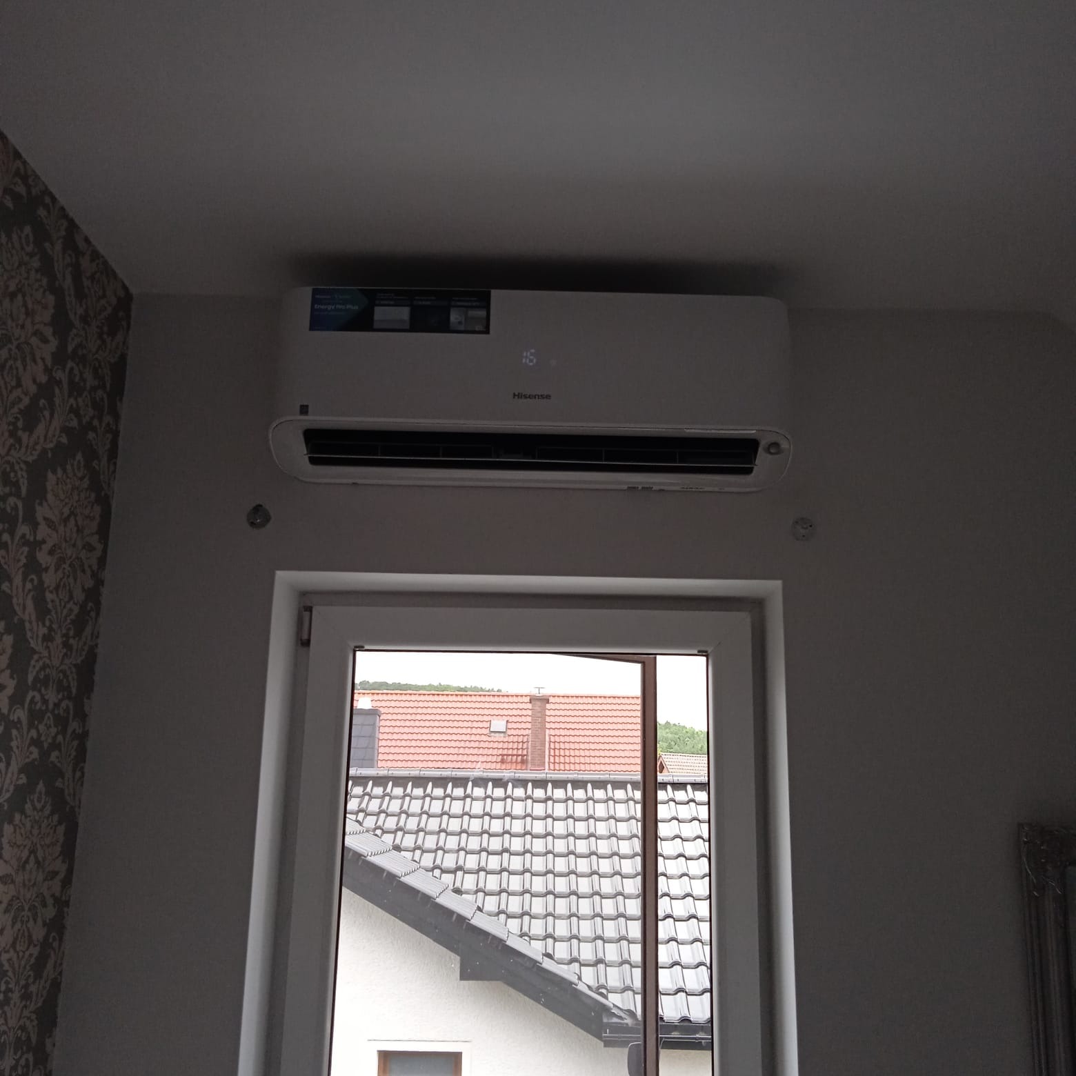 Biała klimatyzacja Hisense zamontowana nad oknem w pokoju z wzorzystą tapetą, widok na dachy domów za oknem.