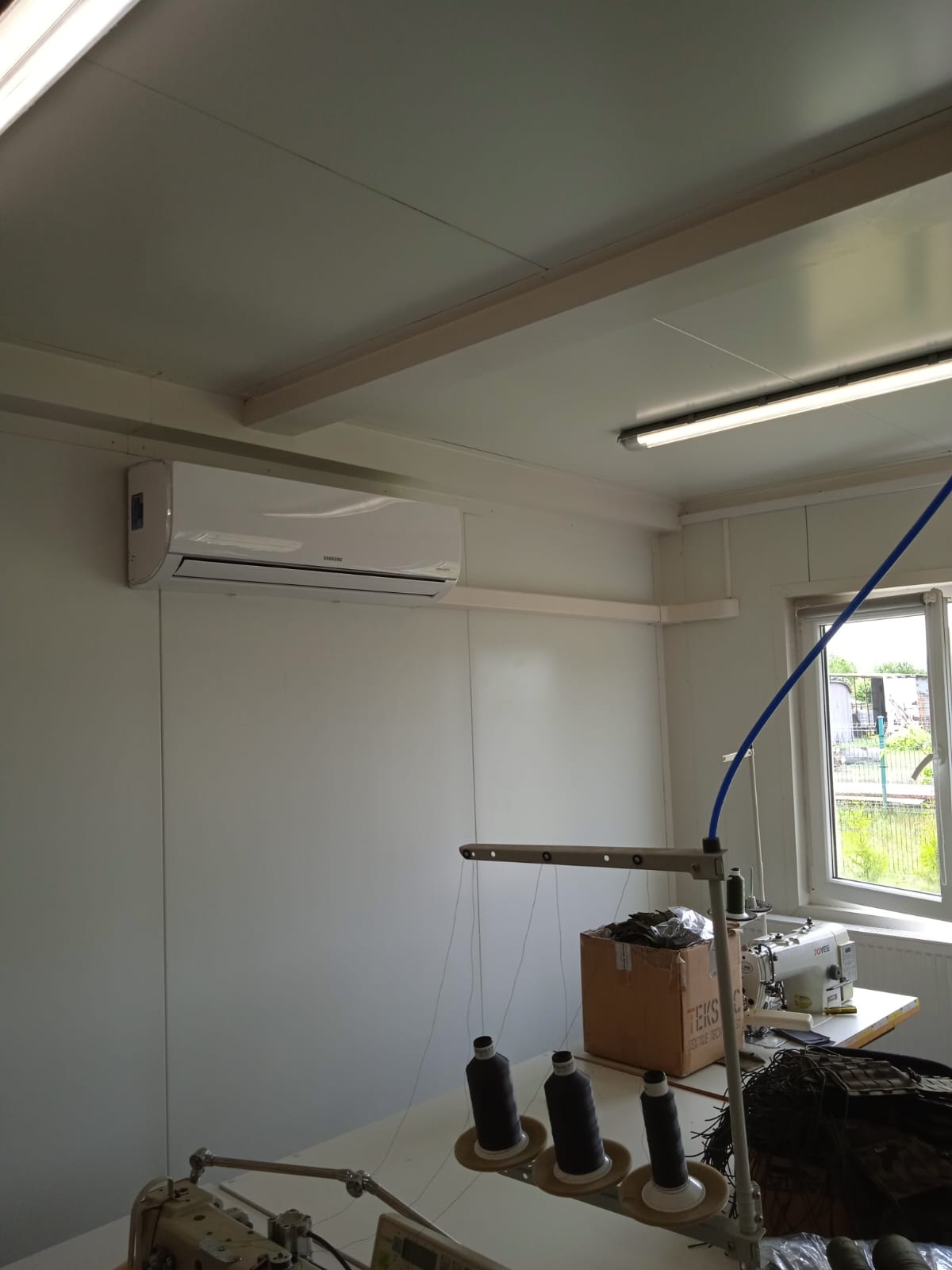 Biały klimatyzator Samsung zamontowany na ścianie w pomieszczeniu produkcyjnym, widoczne maszyny do szycia i nici.