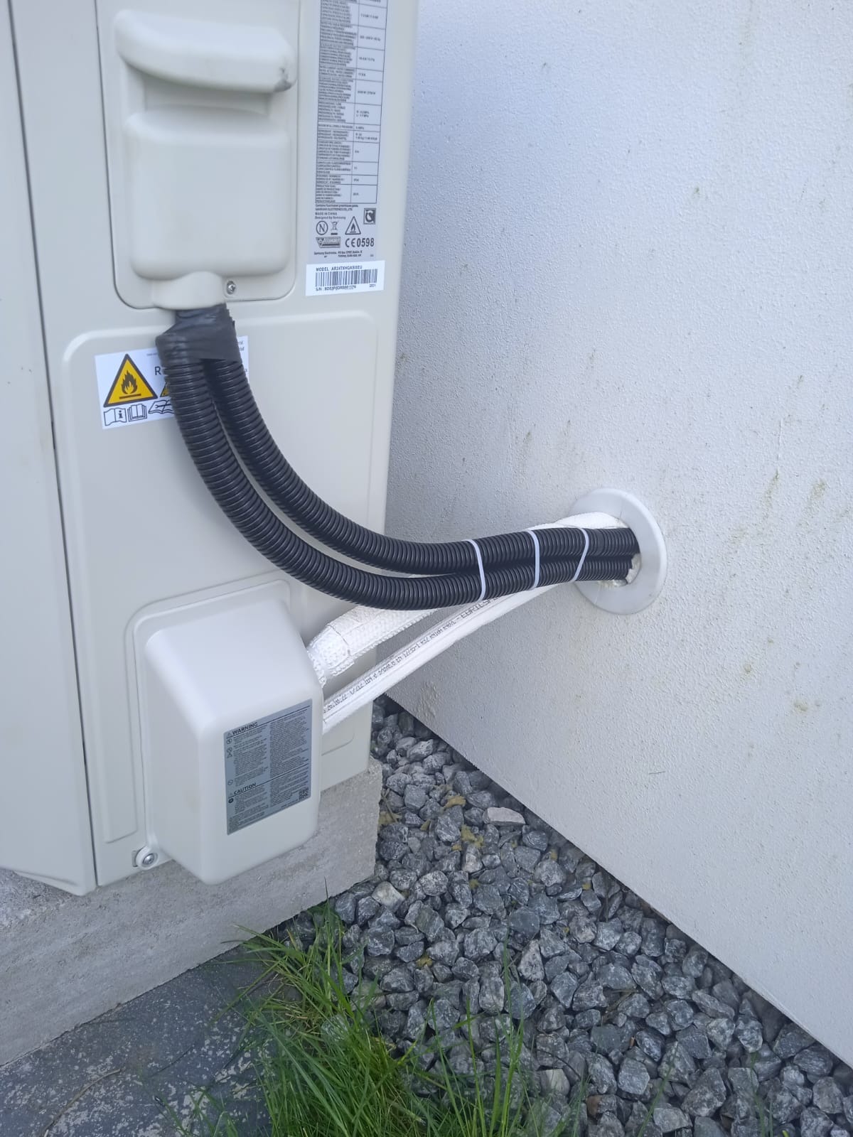 Zewnętrzna jednostka klimatyzacji z podwójnymi rurami odprowadzającymi przez otwór w ścianie, zabezpieczonym kołnierzem, widoczne kamienie i trawa u podstawy.