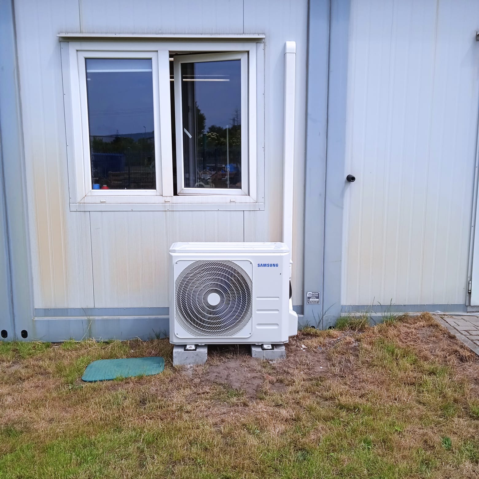 Zewnętrzna jednostka klimatyzacji Samsung zamontowana na betonowych podstawach przy metalowym kontenerze z otwartym oknem, widoczne ślady zabrudzeń na elewacji.