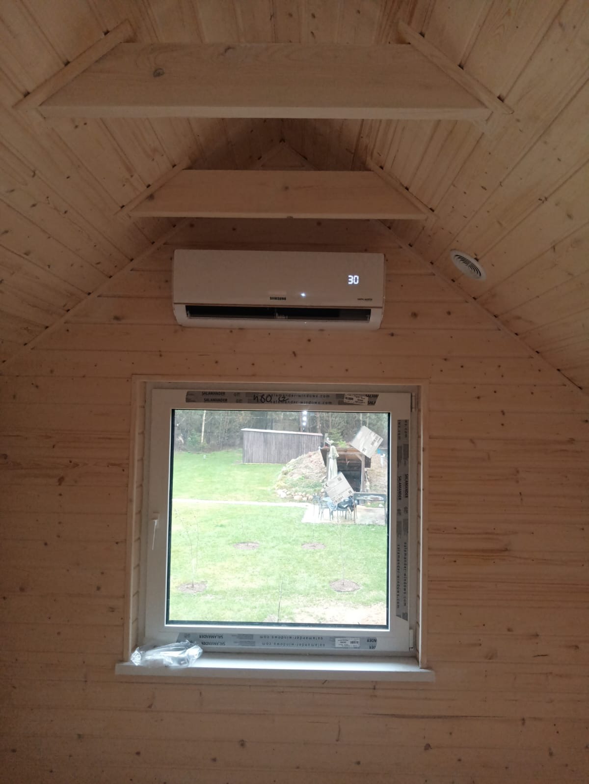 Wnętrze drewnianego domku z zainstalowanym klimatyzatorem Samsung, ustawionym na 30 stopni, widok przez okno na zielony ogród.