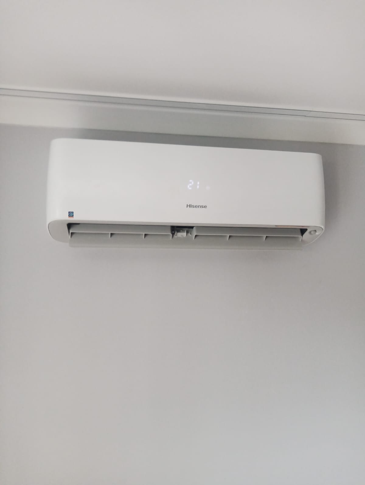 Biała jednostka klimatyzacyjna Hisense zamontowana na jasnoszarej ścianie, wyświetlająca temperaturę 21 stopni.