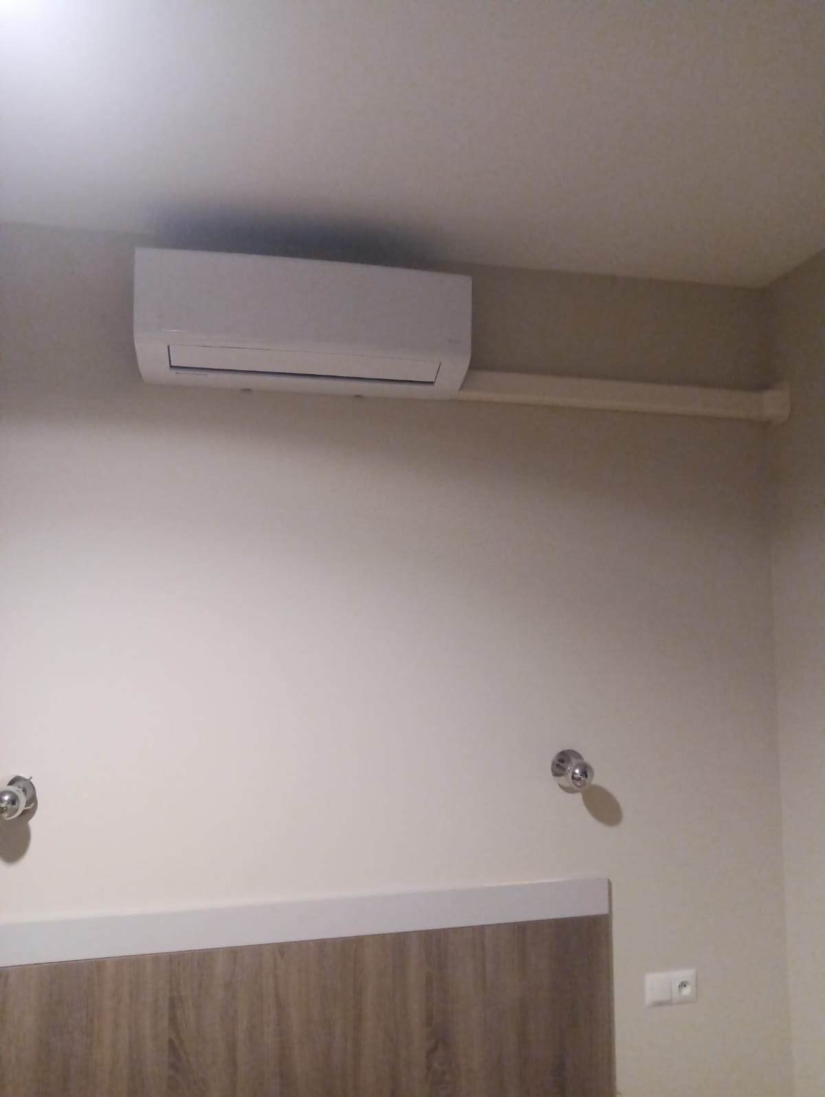 Biały klimatyzator ścienny zamontowany w pomieszczeniu z beżowymi ścianami, widoczna rura instalacyjna, chromowane gałki na ścianie i fragment drewnianej boazerii.