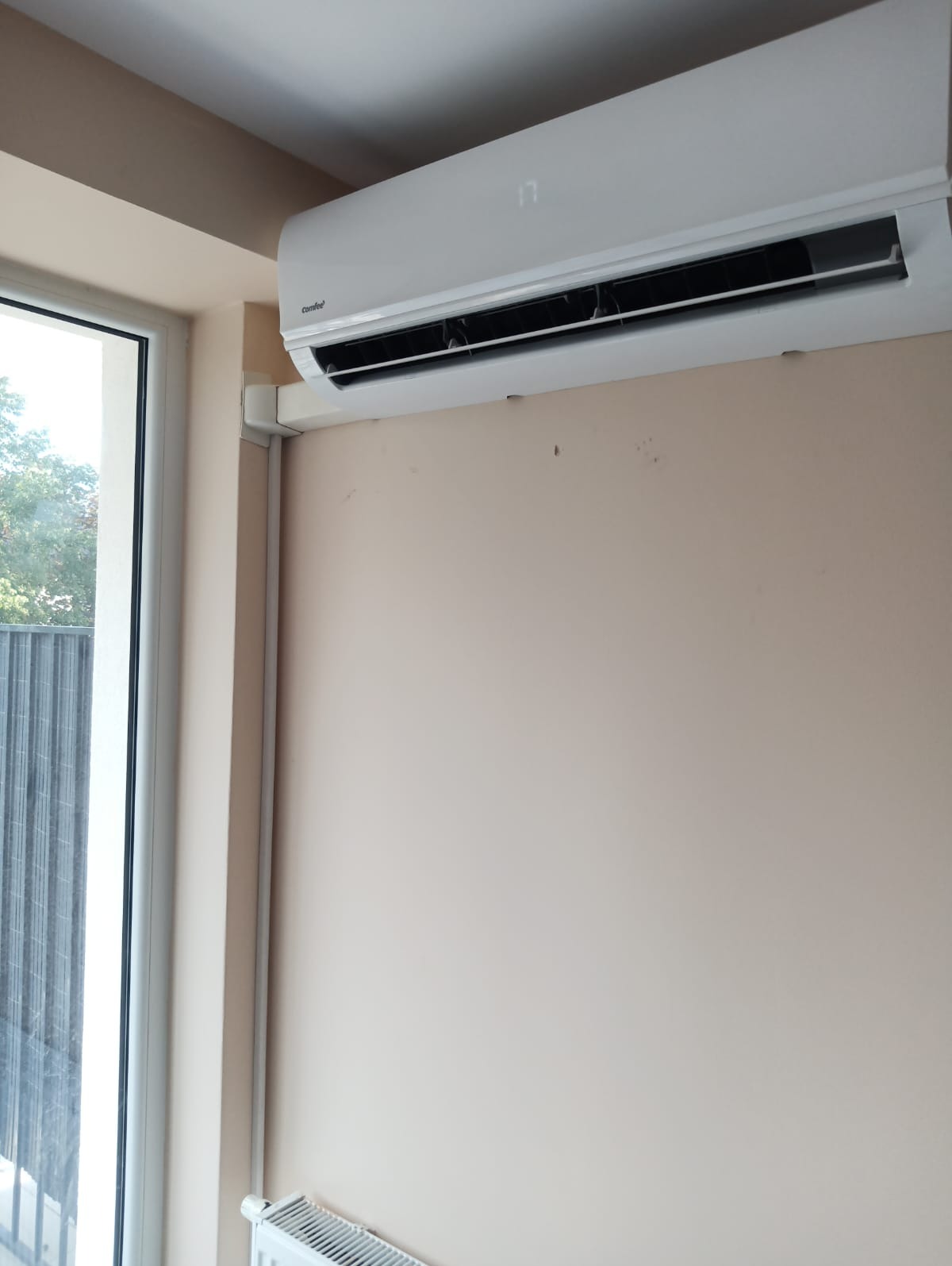 Biały klimatyzator ścienny marki Comfee, ustawiony na 17 stopni, zamontowany w pomieszczeniu z oknem i grzejnikiem, widoczne ślady na ścianie.
