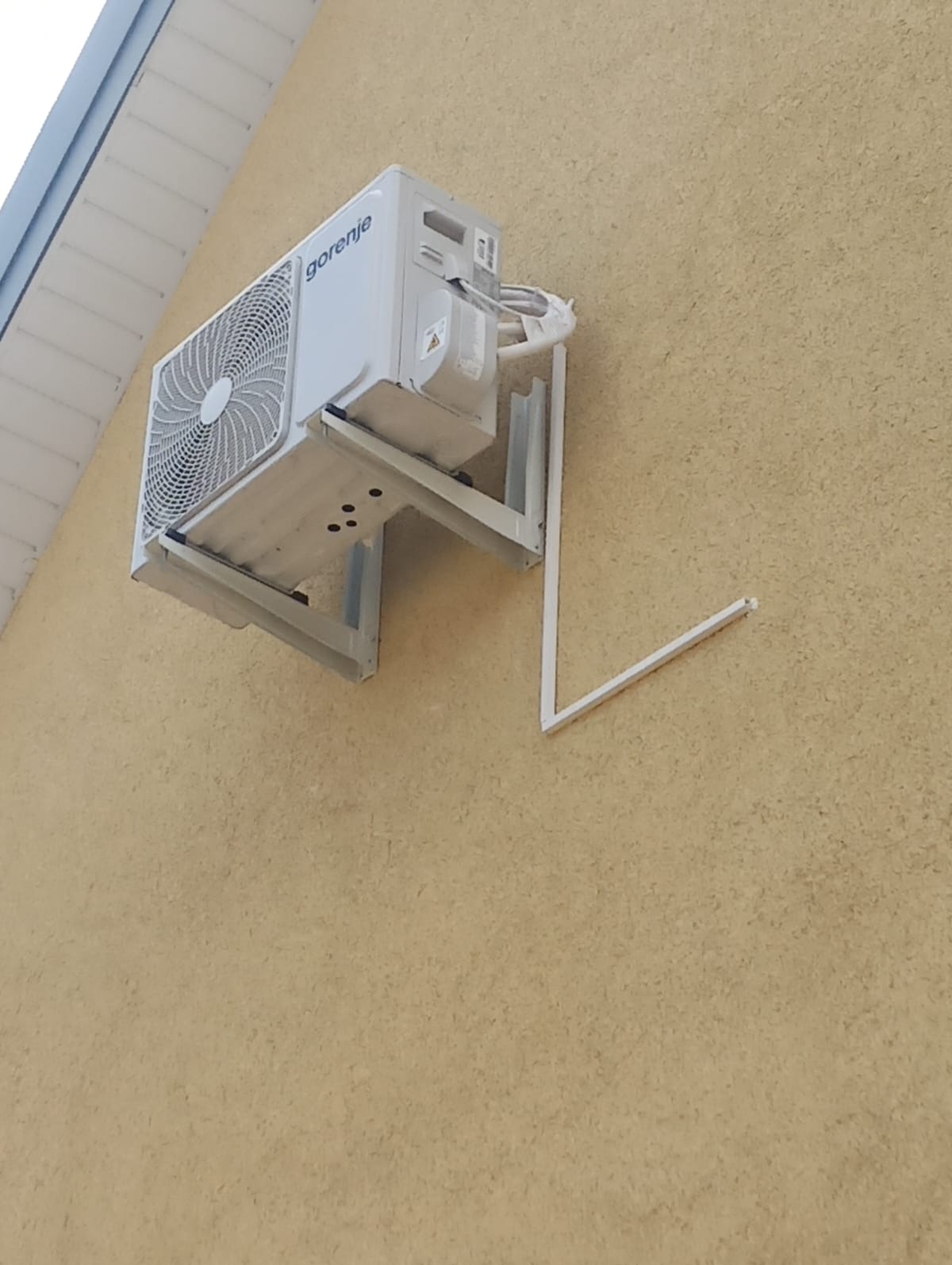 Biała jednostka zewnętrzna klimatyzacji marki Gorenje zamontowana na wsporniku na żółtej, teksturowanej ścianie budynku. Fragment wspornika jest ułamany.