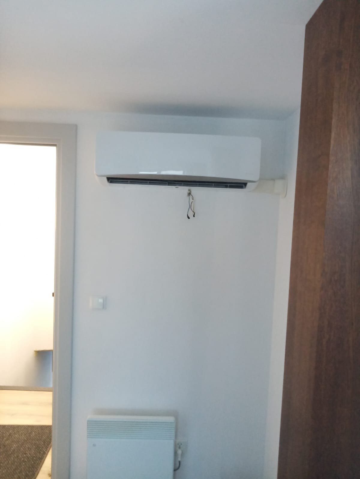 Biały klimatyzator ścienny zamontowany w jasnym pomieszczeniu, z widocznymi przewodami i grzejnikiem elektrycznym poniżej.