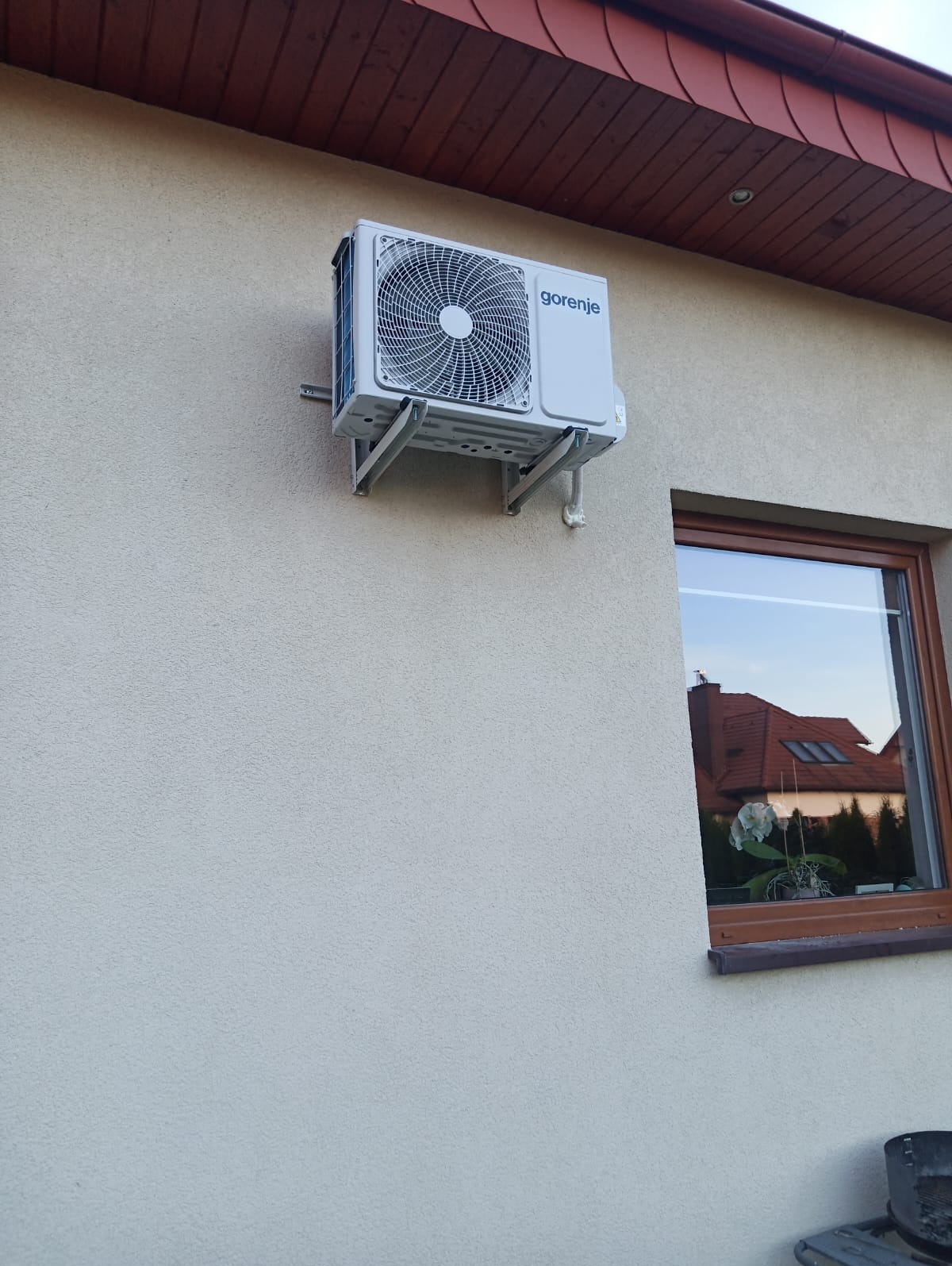Zewnętrzna jednostka klimatyzacji Gorenje zamontowana na elewacji domu obok okna z odbiciem sąsiedniego budynku.