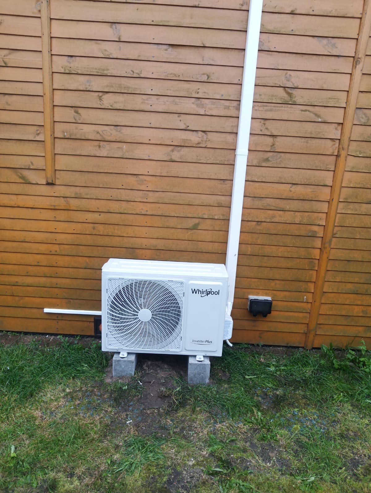 Zewnętrzna jednostka klimatyzacji Whirlpool Inverter Plus zamontowana na betonowych bloczkach przy drewnianej ścianie budynku, z widoczną rurą odprowadzającą.
