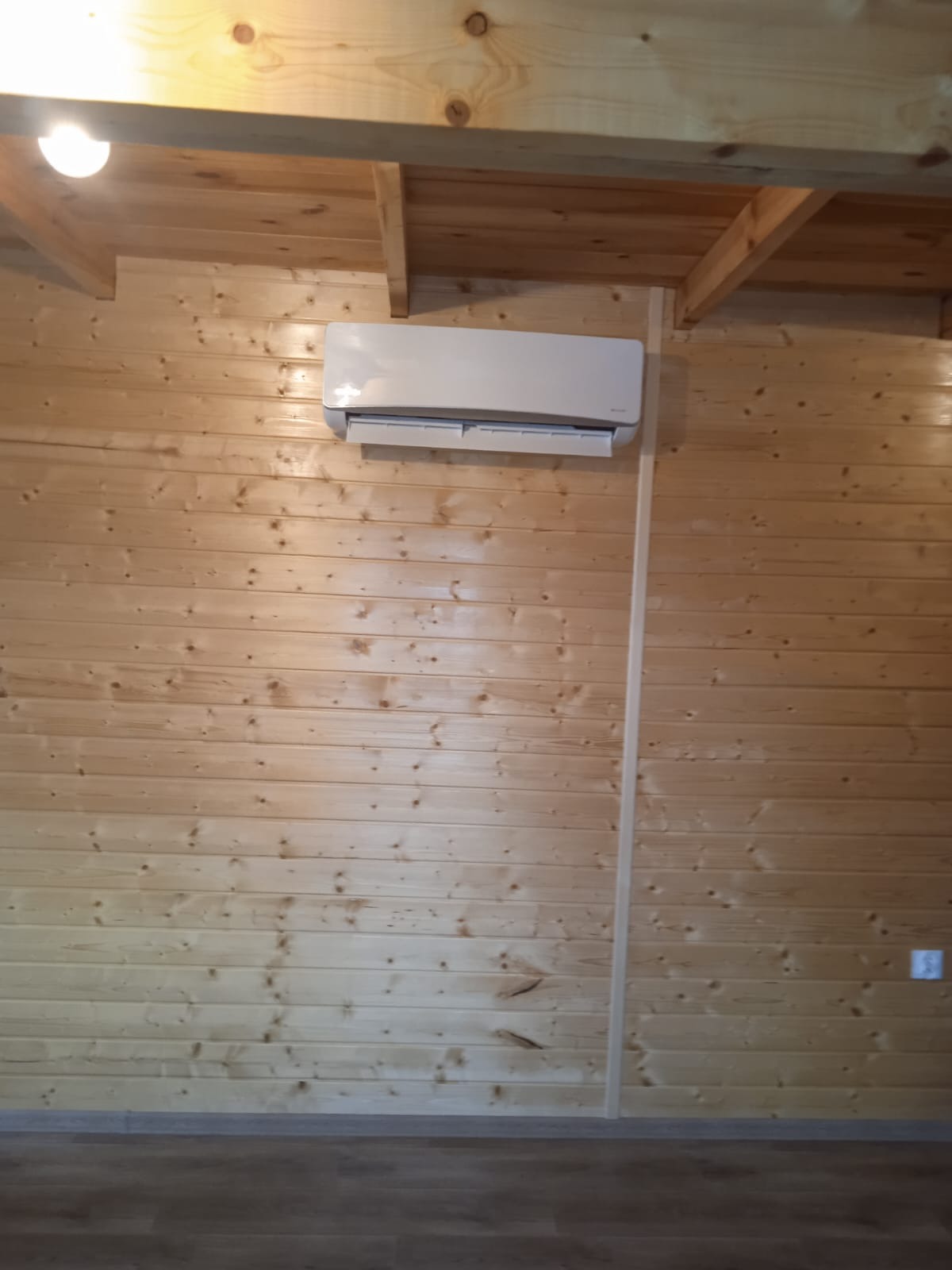 Biały klimatyzator zamontowany na drewnianej ścianie w domu z boazerią, widoczna lampa na suficie.