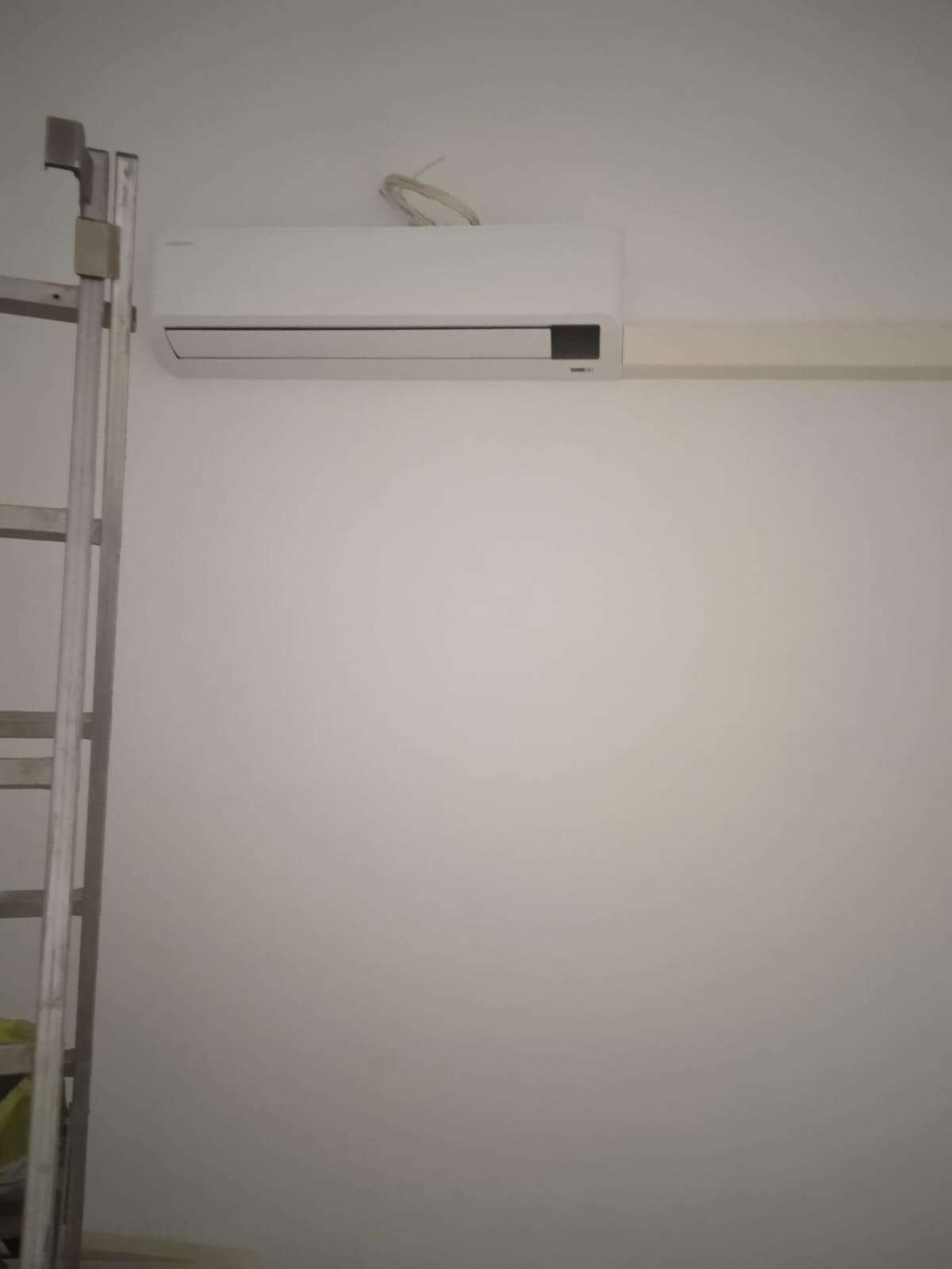 Biała jednostka klimatyzacji zamontowana na ścianie, widoczne przewody i drabina oparta o ścianę.