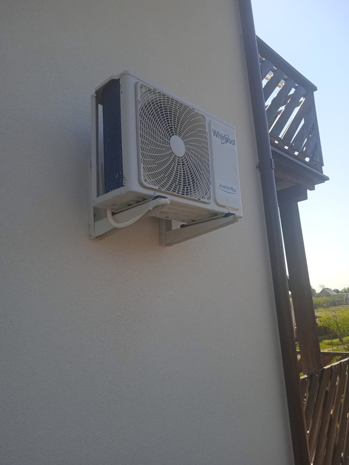 Biała jednostka klimatyzacji Whirlpool Inverter Plus zamontowana na zewnętrznej ścianie budynku, widoczne rury odprowadzające skropliny, w tle fragment drewnianego balkonu i zieleń.