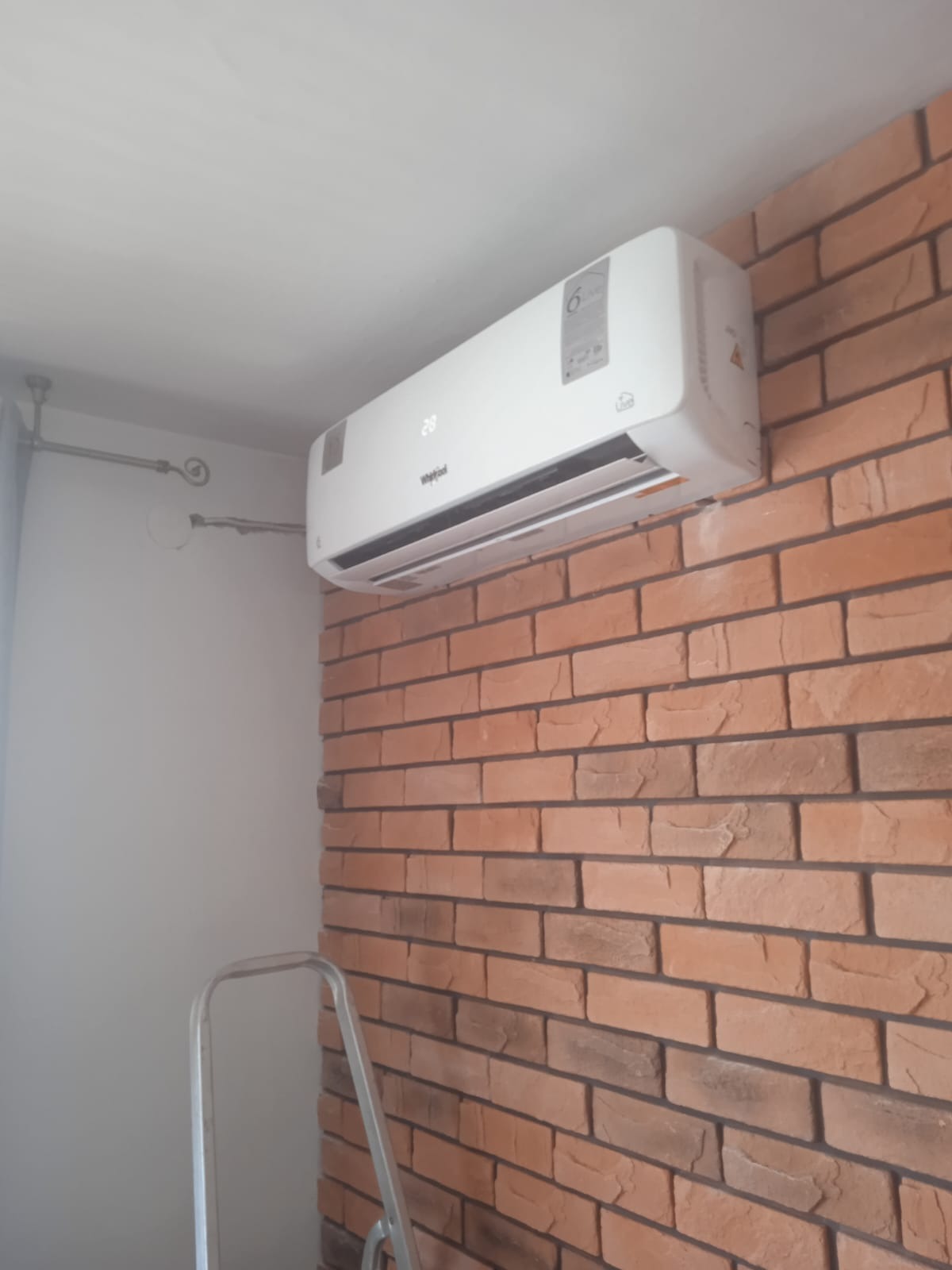 Biały klimatyzator Whirlpool zamontowany na ścianie z cegły, widoczna drabina aluminiowa.