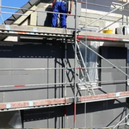 Ocieplanie elewacji domu jednorodzinnego szarym styropianem, widoczne rusztowania i pracownik w niebieskim kombinezonie na górnym poziomie rusztowania.