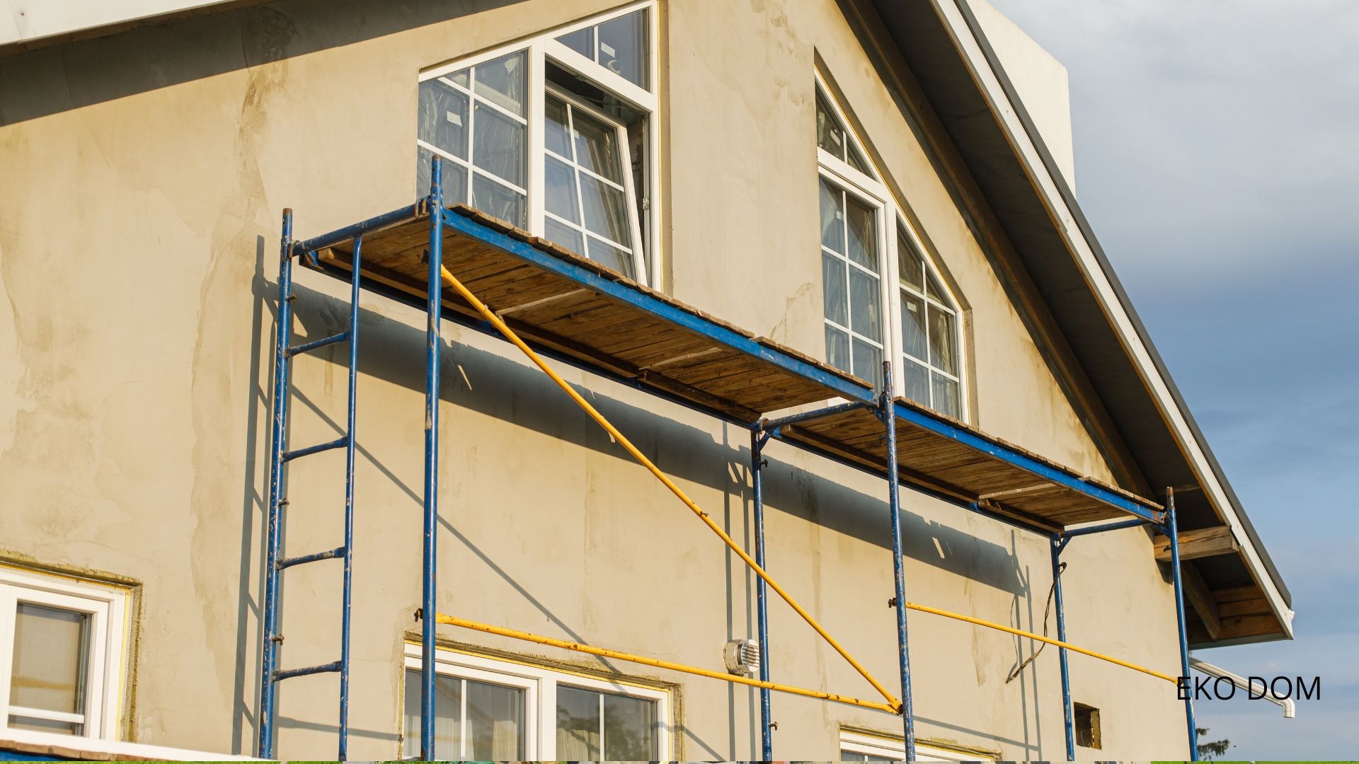Ściana domu w trakcie prac elewacyjnych, widoczne rusztowanie, okna z białymi ramami, elewacja w odcieniach beżu przed nałożeniem warstwy wykończeniowej.