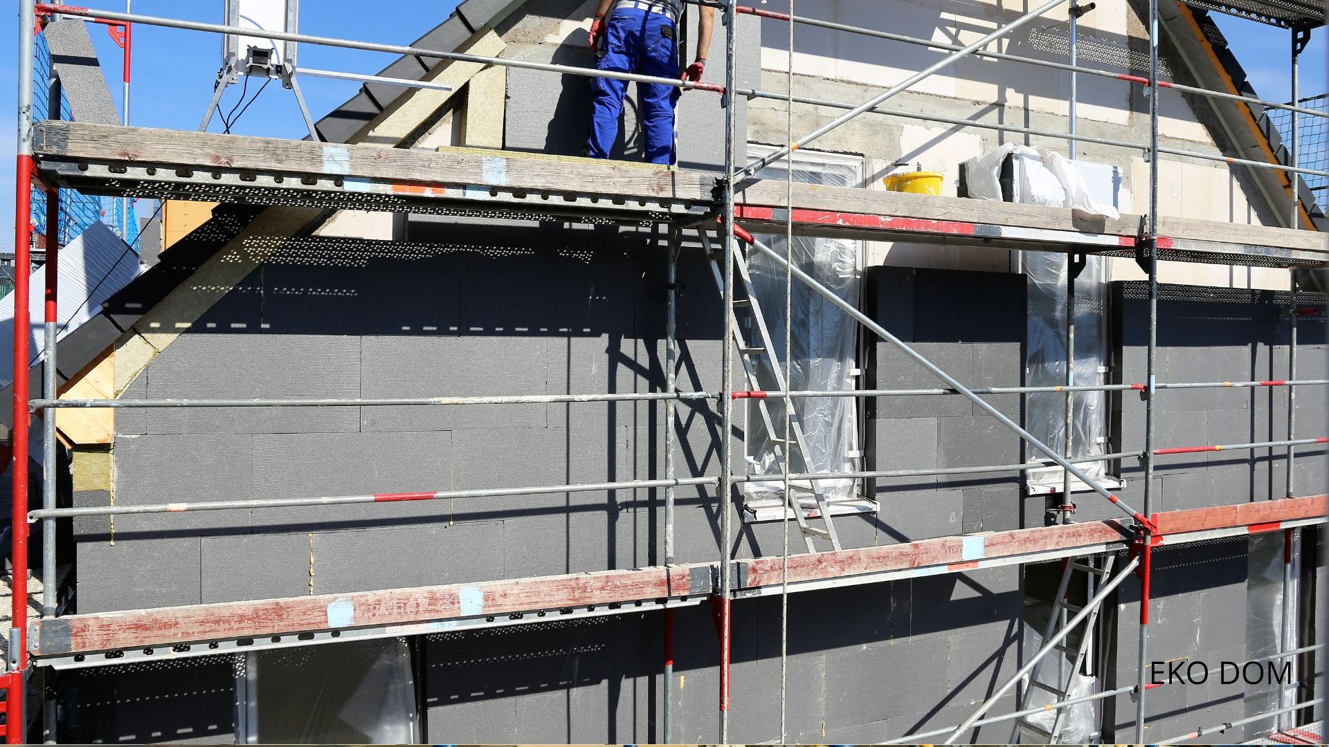 Ocieplanie elewacji domu jednorodzinnego szarym styropianem, widoczne rusztowania i pracownik w niebieskim kombinezonie na górnym poziomie rusztowania.