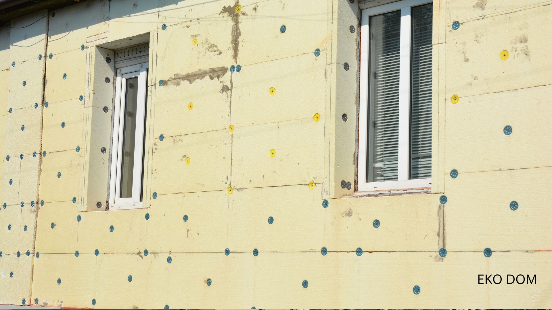 Ocieplona ściana budynku z dwoma oknami, widoczne kołki mocujące styropian z niebieskimi i żółtymi elementami, na dole widoczny napis EKO DOM.