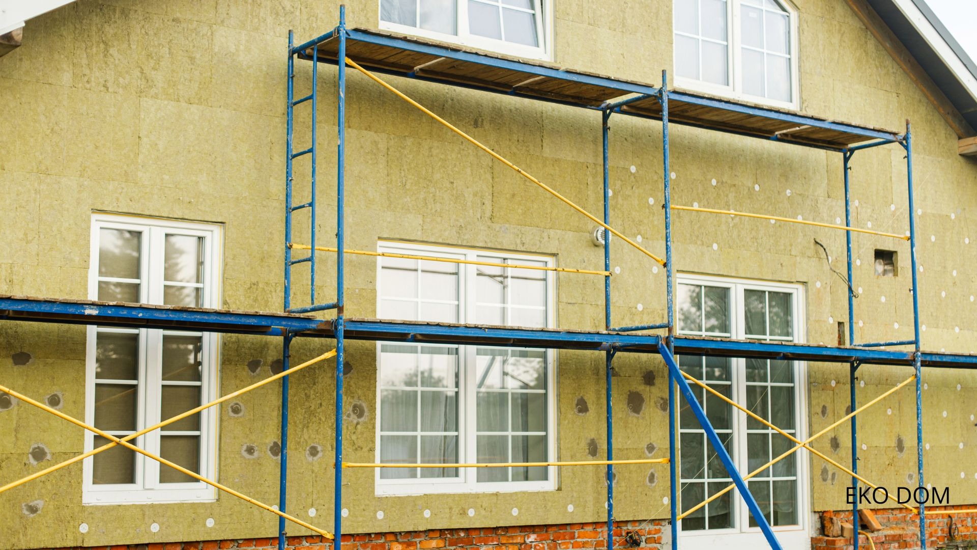 Dom w trakcie ocieplania, widoczne rusztowanie, warstwa izolacyjna z wełny mineralnej i kołki mocujące, dolna część ściany z cegły, okna z białymi ramami.