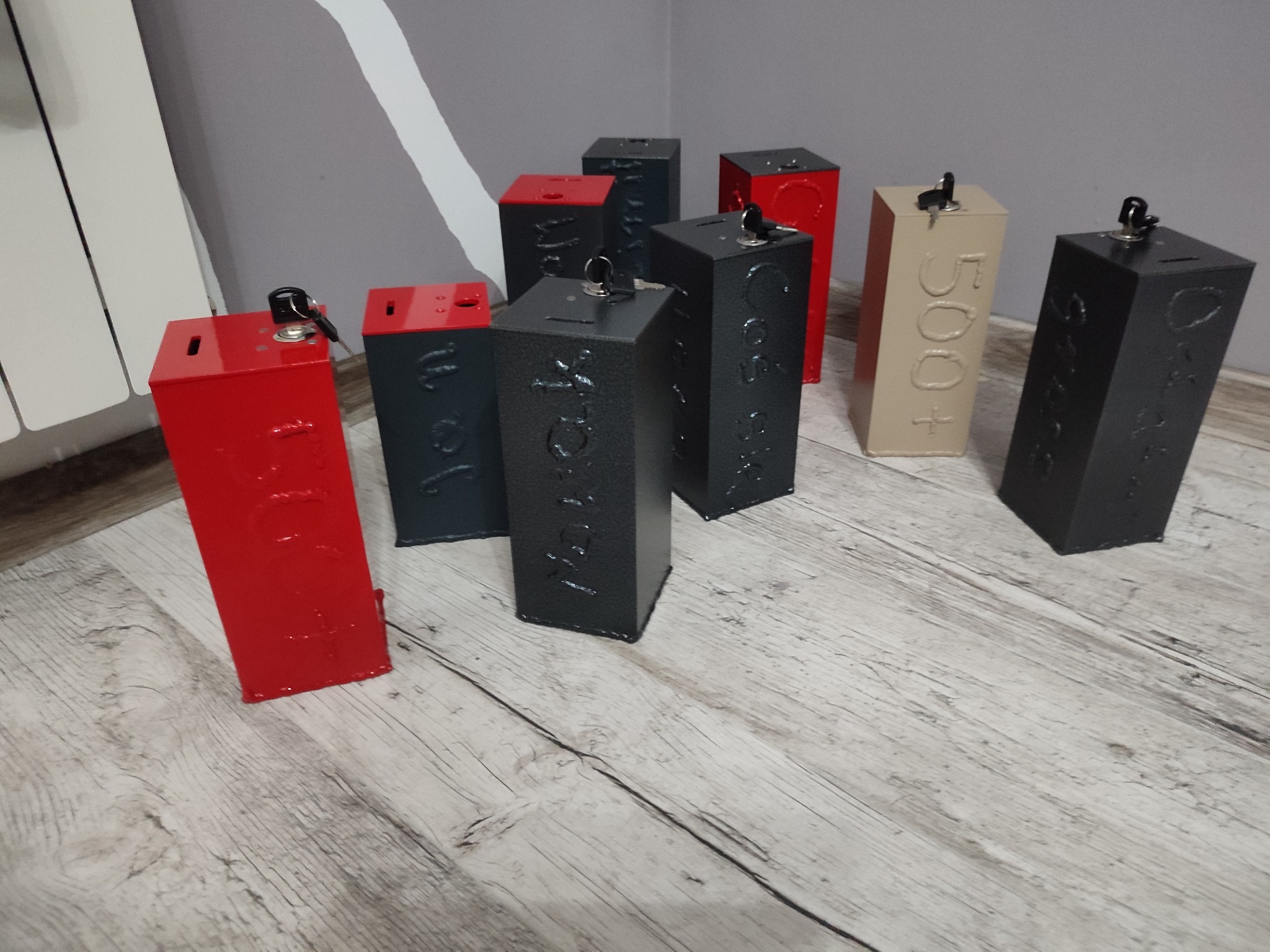 Kolekcja metalowych, prostopadłościennych skarbonek zamykanych na klucz, w kolorach czerwonym, czarnym i beżowym z napisem '500+' na jednej z nich, ustawionych na drewnianej podłodze.