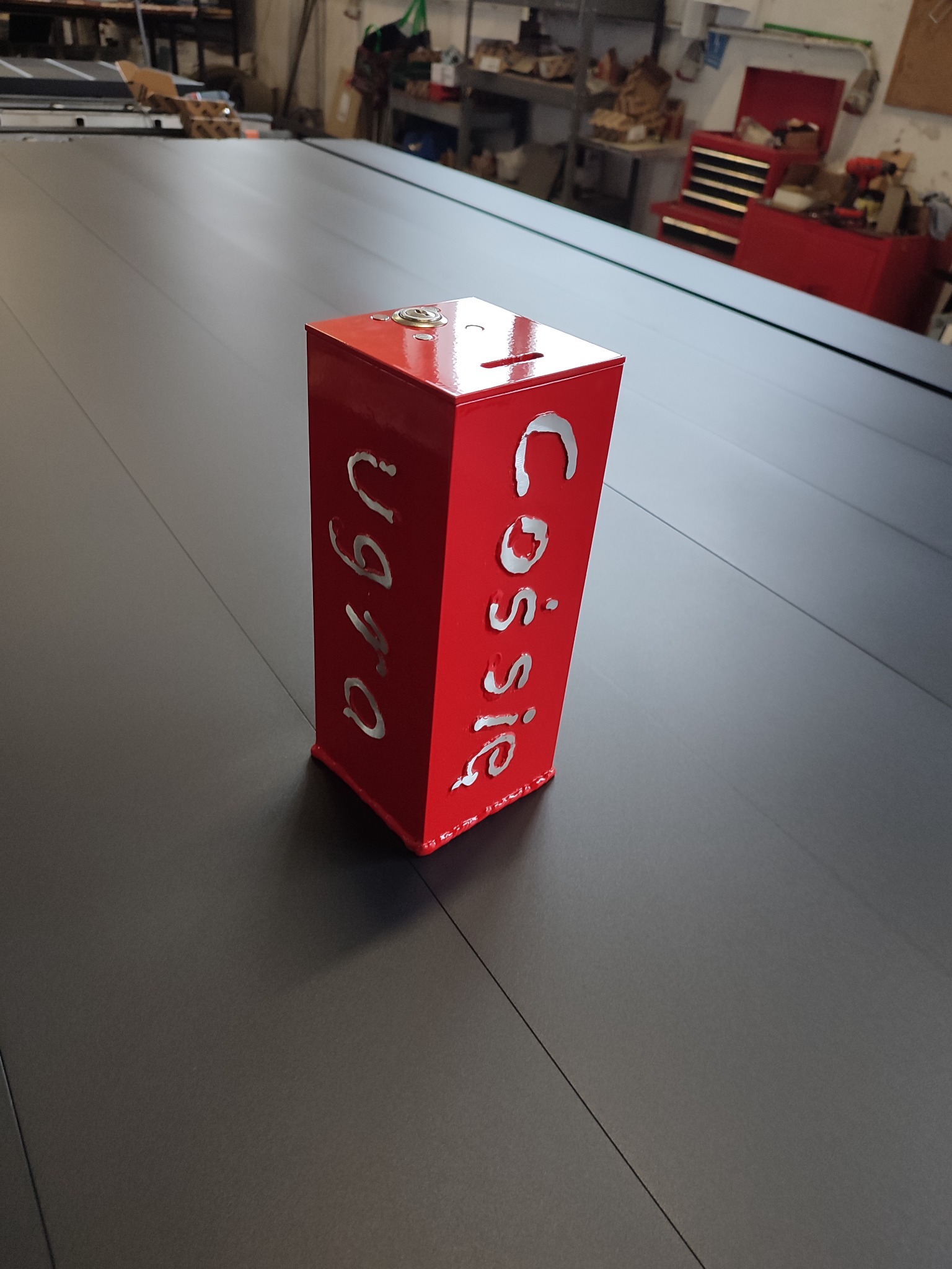 Czerwona, metalowa puszka z wyciętymi laserowo napisami 'Cossie' i 'Ugio' stoi na ciemnej, metalowej powierzchni w warsztacie.