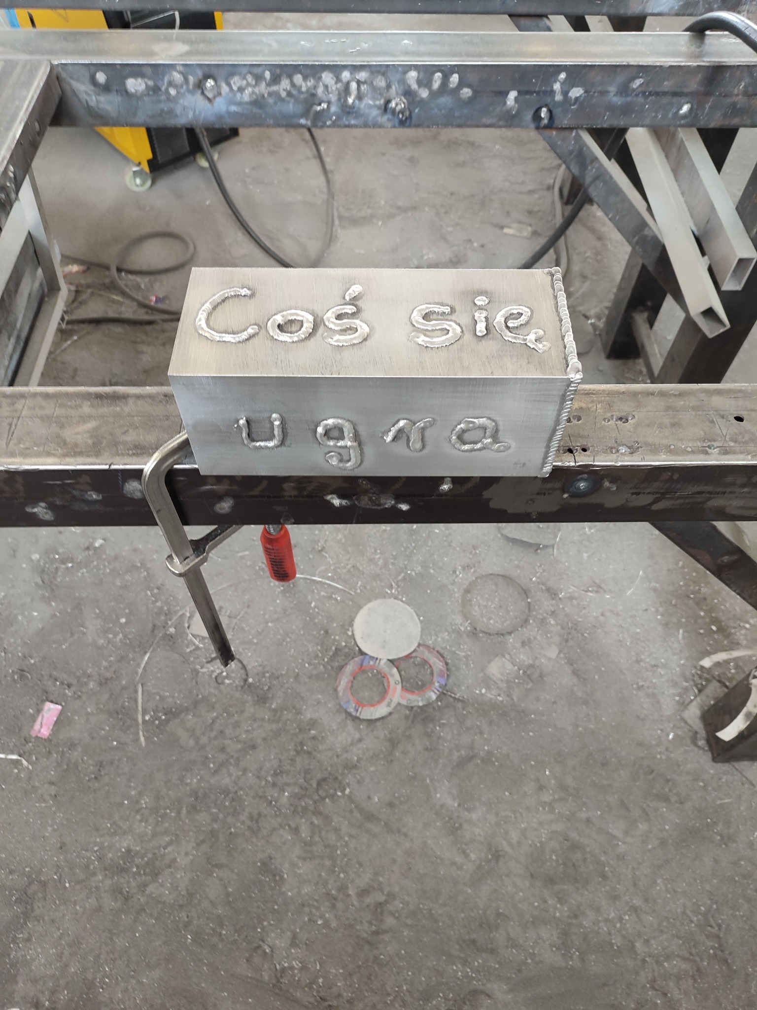 Metalowy prostopadłościan z napisem 'Coś się ugra' wykonanym metodą spawania, przymocowany ściskiem stolarskim do metalowej konstrukcji.