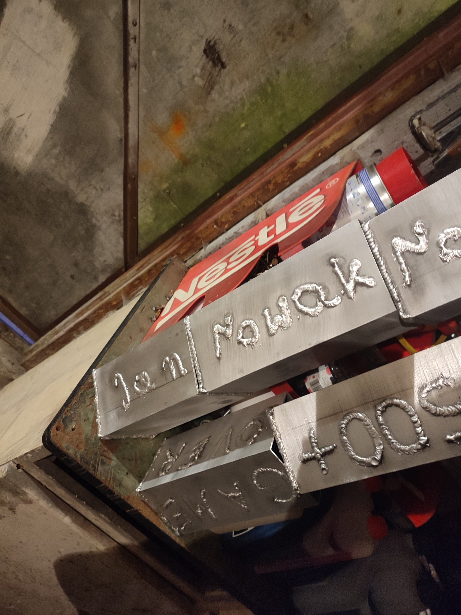 Metalowe profile z napisem 'Jan Nowak' oraz 'BMW +100', wykonane techniką spawania, widoczne w warsztatowym otoczeniu z elementami reklamowymi w tle.