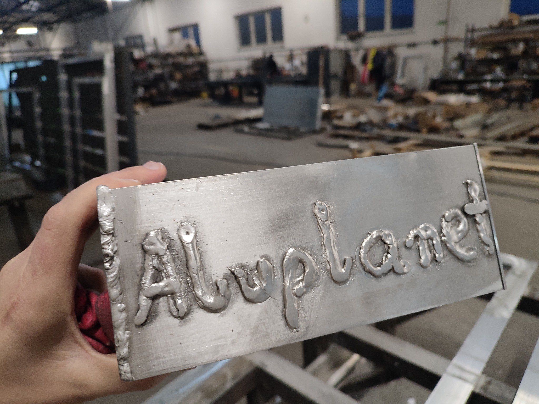Metalowy blok z napisem 'Allplanet' wykonanym spoinami, trzymany w dłoni na tle hali produkcyjnej.