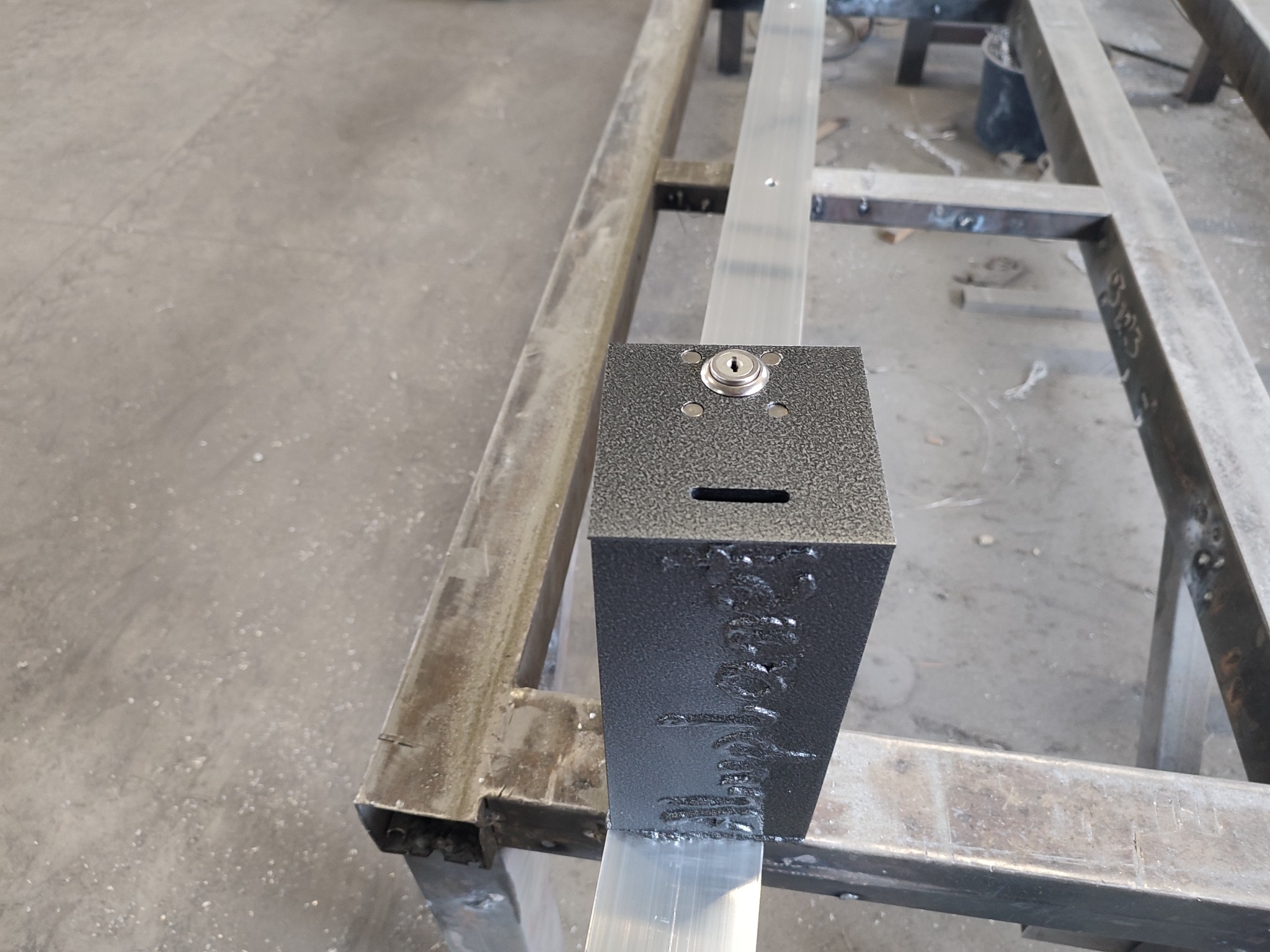 Metalowa skrzynka z zamkiem i otworem wrzutowym przymocowana do aluminiowego profilu na metalowej konstrukcji, widoczne ślady spawania.