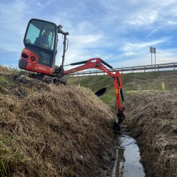 Piotr Żubert - Czerwona mini koparka Kubota IX019-4 w trakcie kopania wąskiego rowu melioracyjnego na zarośniętym terenie, widok z boku, pochmurne niebo.