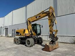 Żółta koparka kołowa CAT z łyżką, zaparkowana na placu budowy przed szarą halą. Widoczne opony z bieżnikiem i ramię koparki wzniesione do góry.