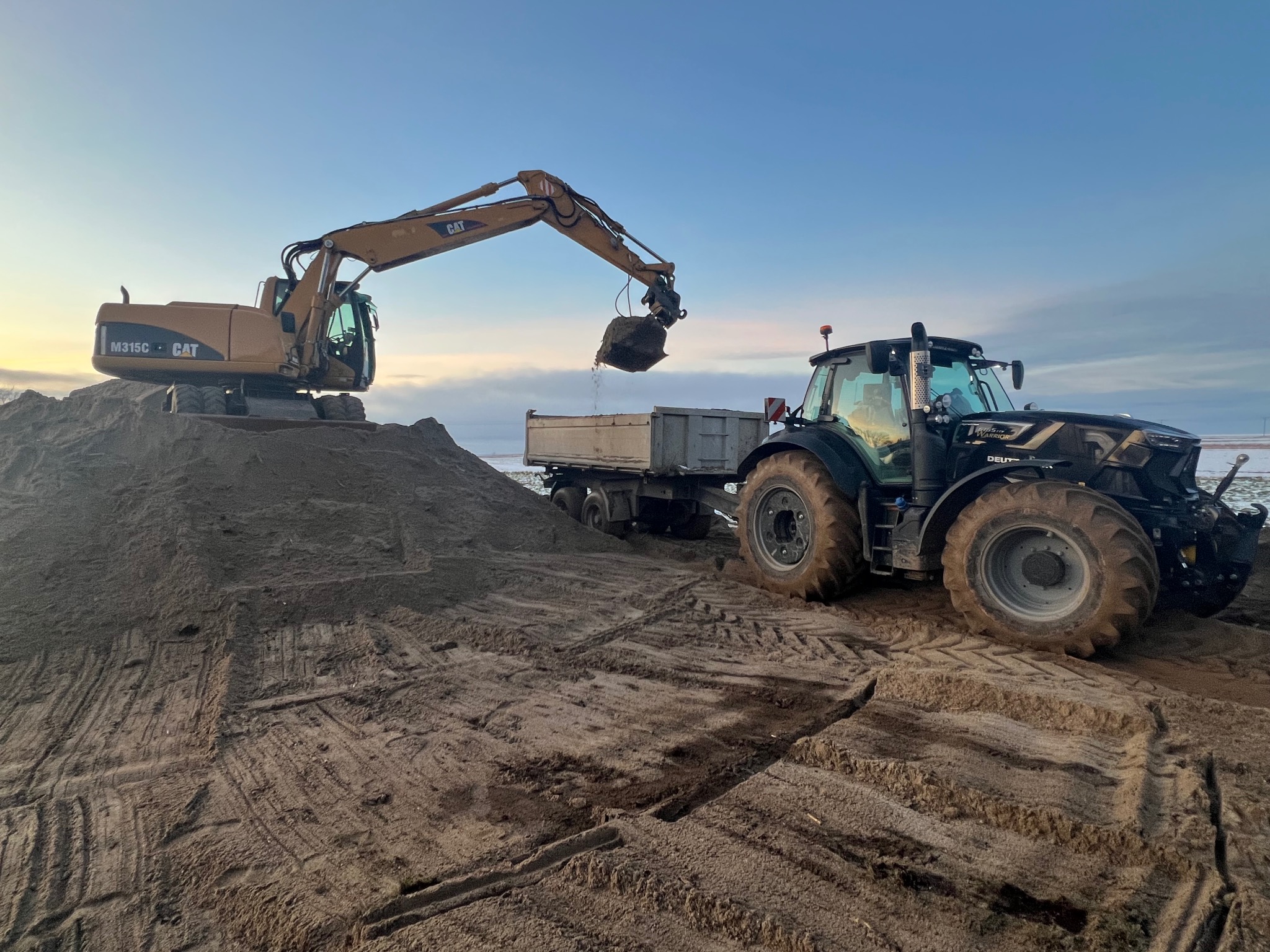 Żółta koparka CAT nabiera ziemię z hałdy i wysypuje ją do przyczepy ciągnika Deutz-Fahr Warrior na tle błękitnego nieba. Ślady opon na ziemi.