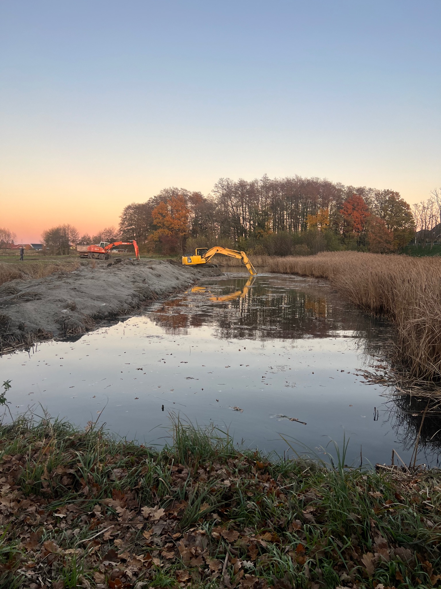 Żółta koparka w trakcie prac ziemnych przy zbiorniku wodnym, odbicie w tafli wody, w tle druga maszyna i drzewa o jesiennych barwach, pomarańczowe niebo o zmierzchu.