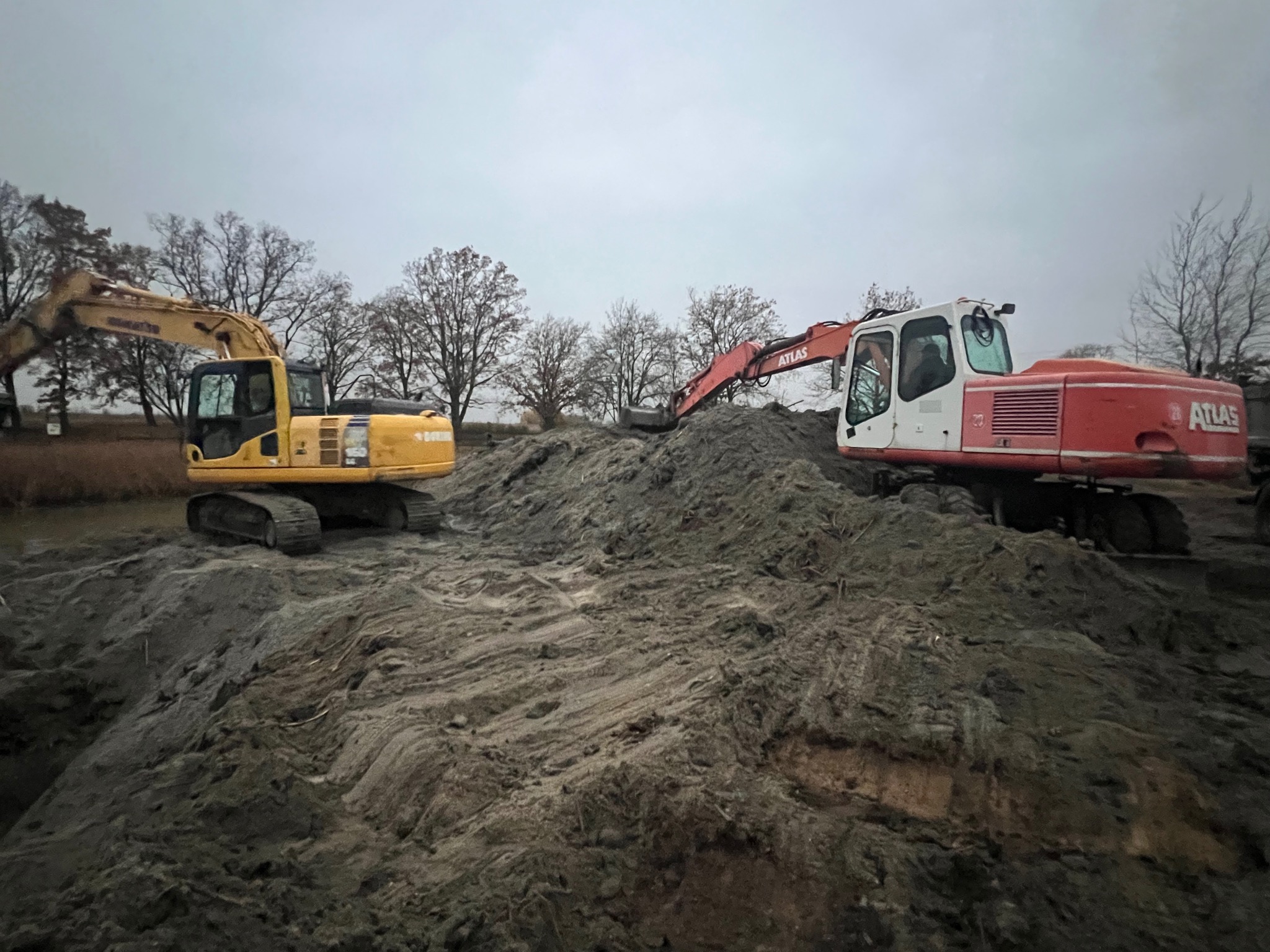 Dwa pracujące, ciężkie, żółto-czarny i czerwono-biały,  ekskawatory Atlas na placu budowy, na tle drzew i pochmurnego nieba. Widoczne świeże pryzmy ziemi.