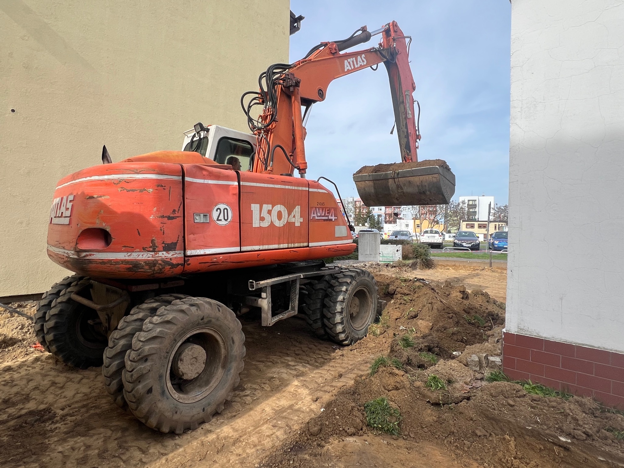Pomarańczowa koparka kołowa Atlas 1504 z pełną łyżką ziemi na placu budowy, widoczna część budynku w tle. Maszyna w trakcie pracy.