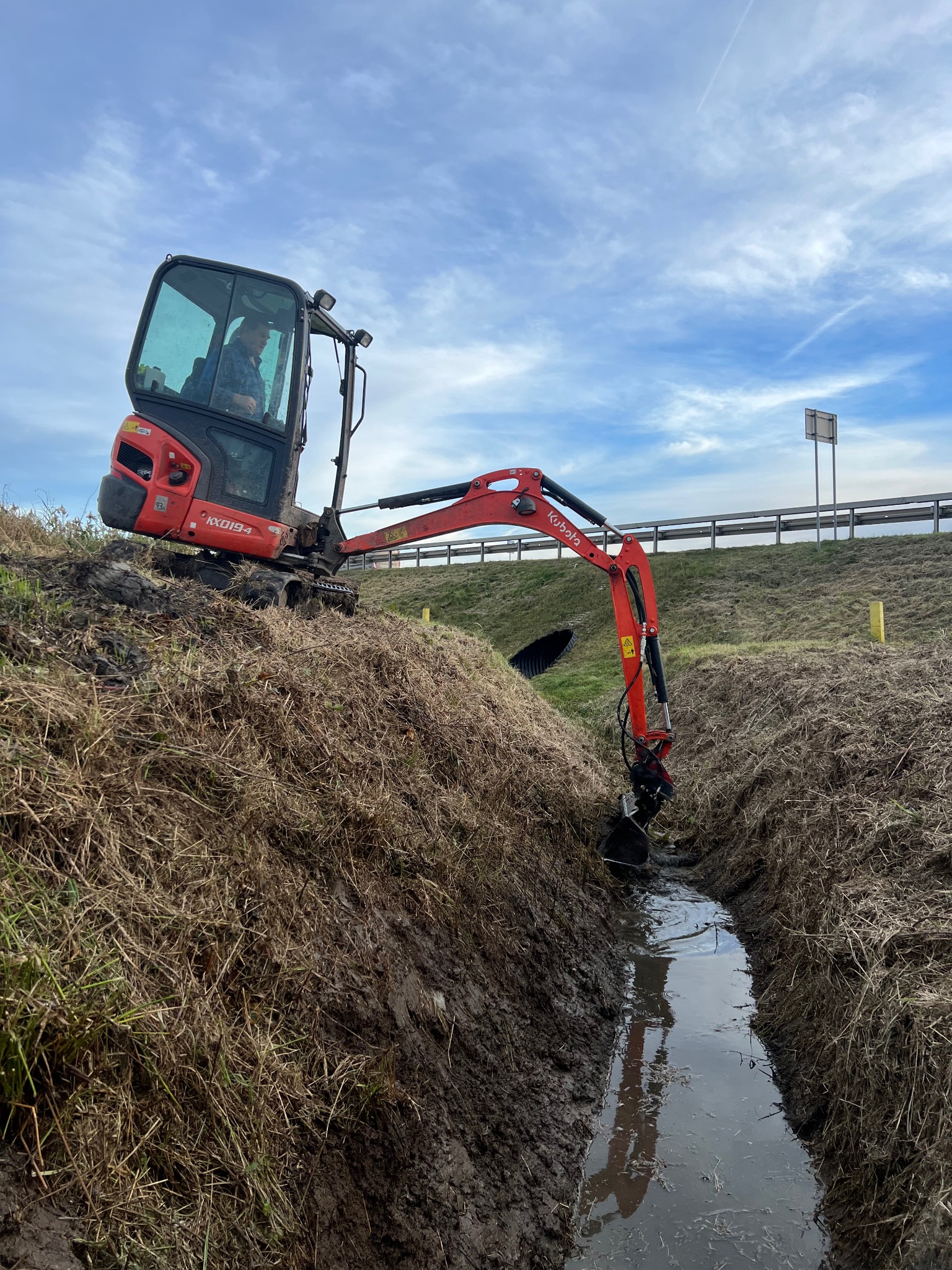 Czerwona mini koparka Kubota IX019-4 w trakcie kopania wąskiego rowu melioracyjnego na zarośniętym terenie, widok z boku, pochmurne niebo.