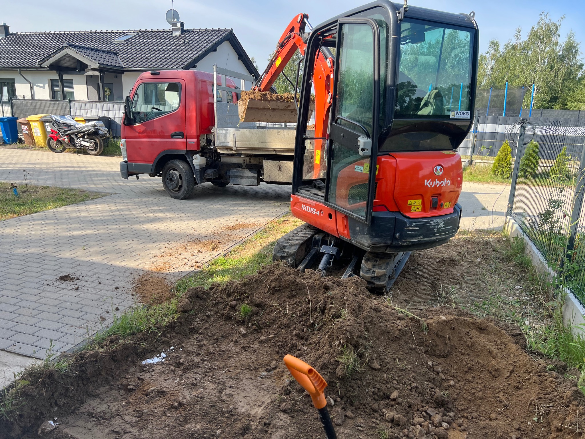 Czerwony mini-koparka Kubota KX019-4 nabiera ziemię do wywrotki na tle domu z szarą dachówką i trampoliny. W dole widoczny fragment wykopu i łopata.