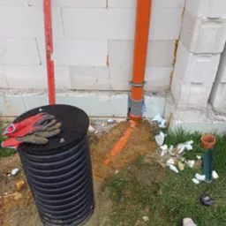 Czarna studzienka kanalizacyjna z pokrywą i roboczymi rękawicami na wierzchu, pomarańczowa rura kanalizacyjna przy ścianie z bloczków, poziomica i szlifierka kątowa w trawie.