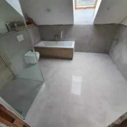 Wnętrze łazienki na poddaszu z oknem dachowym, wyłożonej szarymi płytkami, z kabiną prysznicową ze szkła, zabudowaną wanną z drewnianym frontem i podwieszaną toaletą.