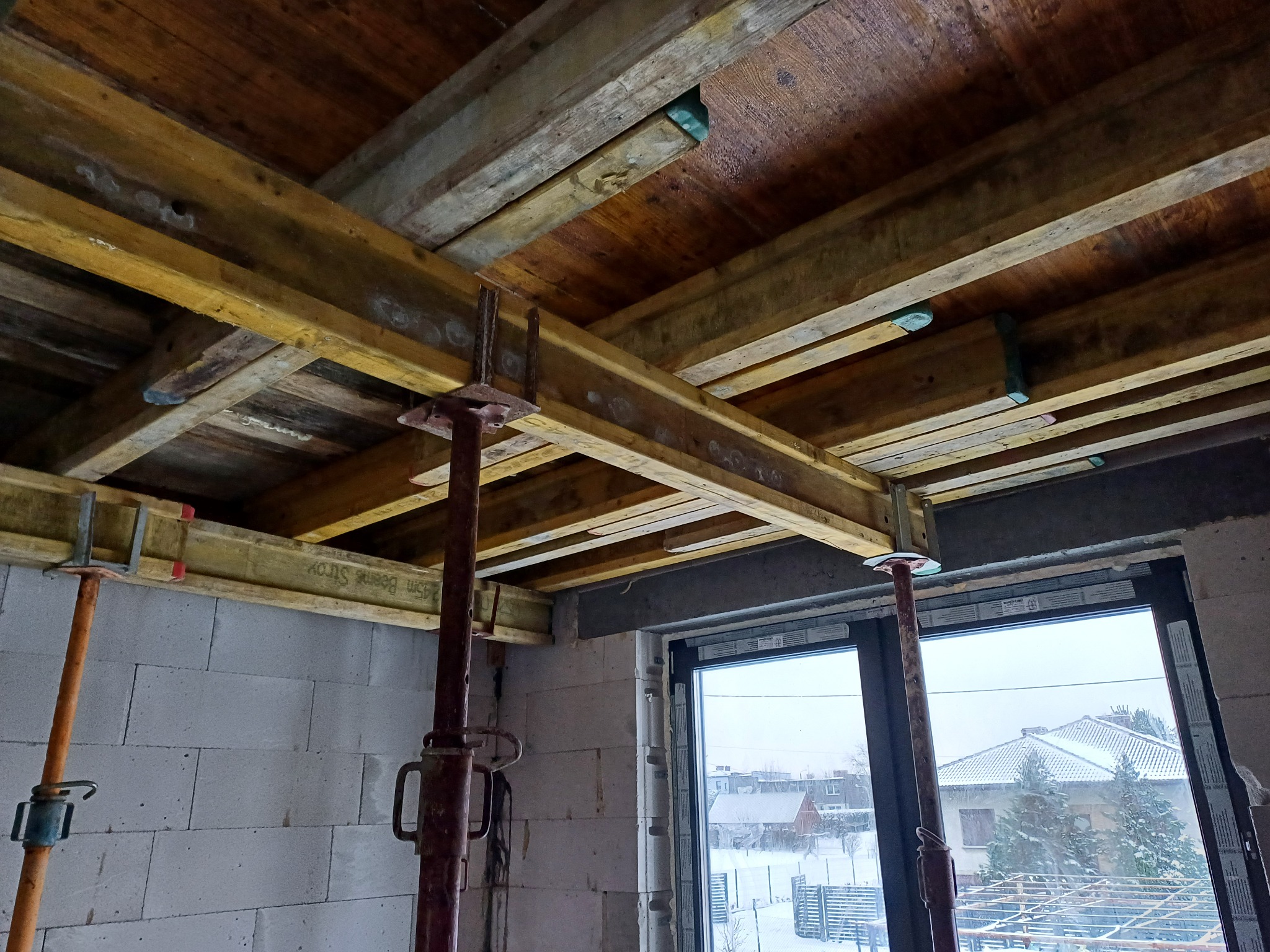 Szczegółowy widok szalunku stropu z drewnianych belek i metalowych podpór w surowym wnętrzu budynku w trakcie budowy, widoczne bloczki z betonu komórkowego i okno z widokiem na zimowy krajobraz.