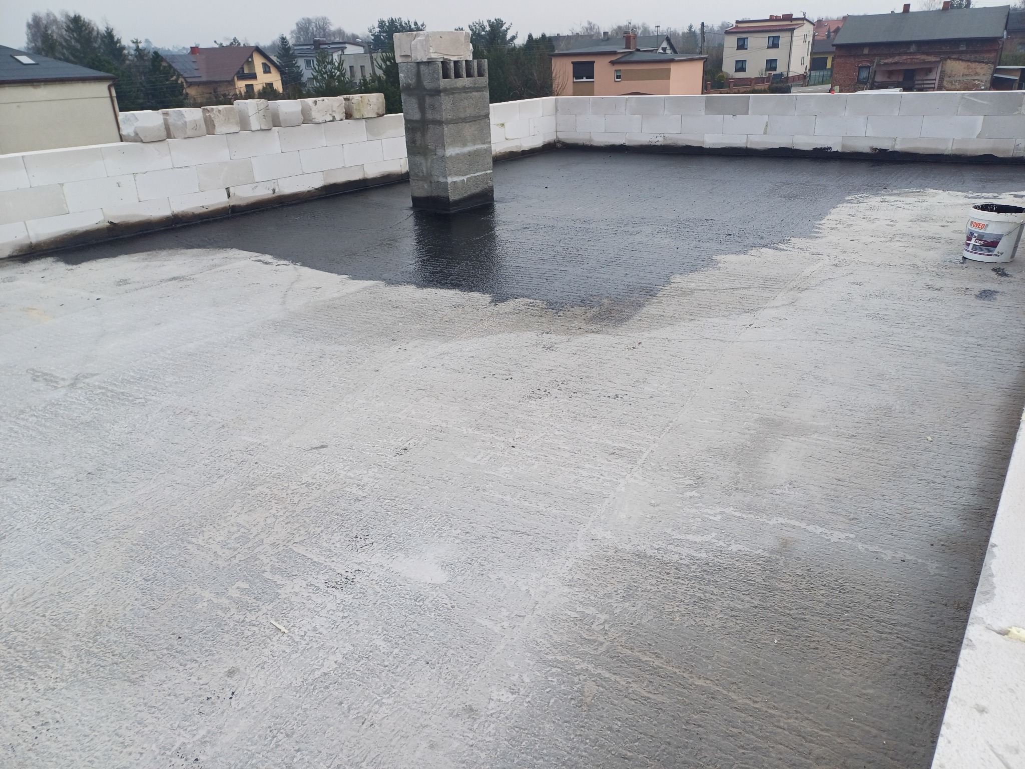 Płaski dach pokryty warstwą hydroizolacyjną, widoczny komin z szarego betonu i białe bloczki na krawędzi dachu, w tle zabudowa jednorodzinna.