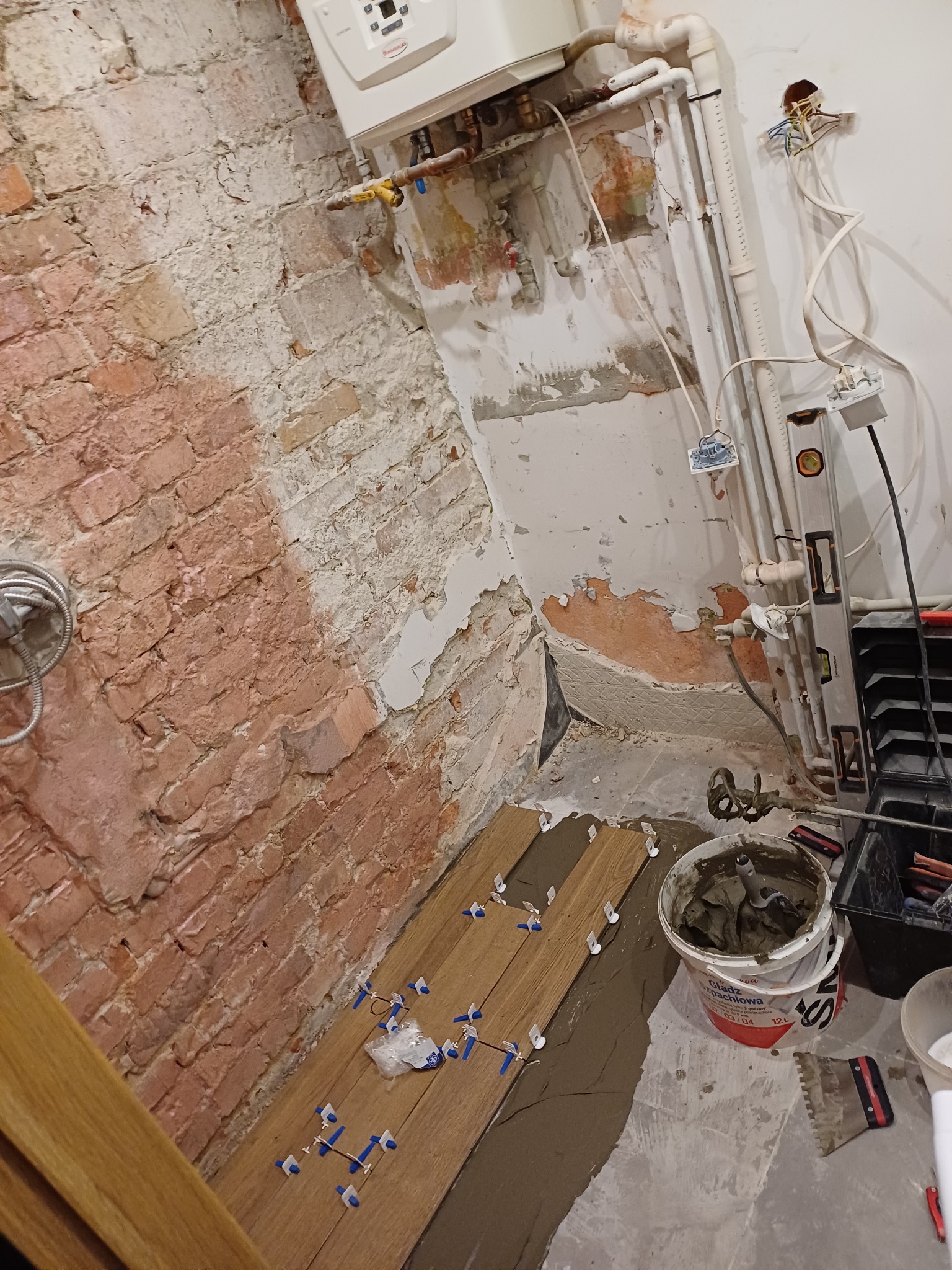 Układanie drewnopodobnych płytek podłogowych w łazience z widoczną starą ceglaną ścianą, grzejnikiem i instalacją hydrauliczną w trakcie remontu.