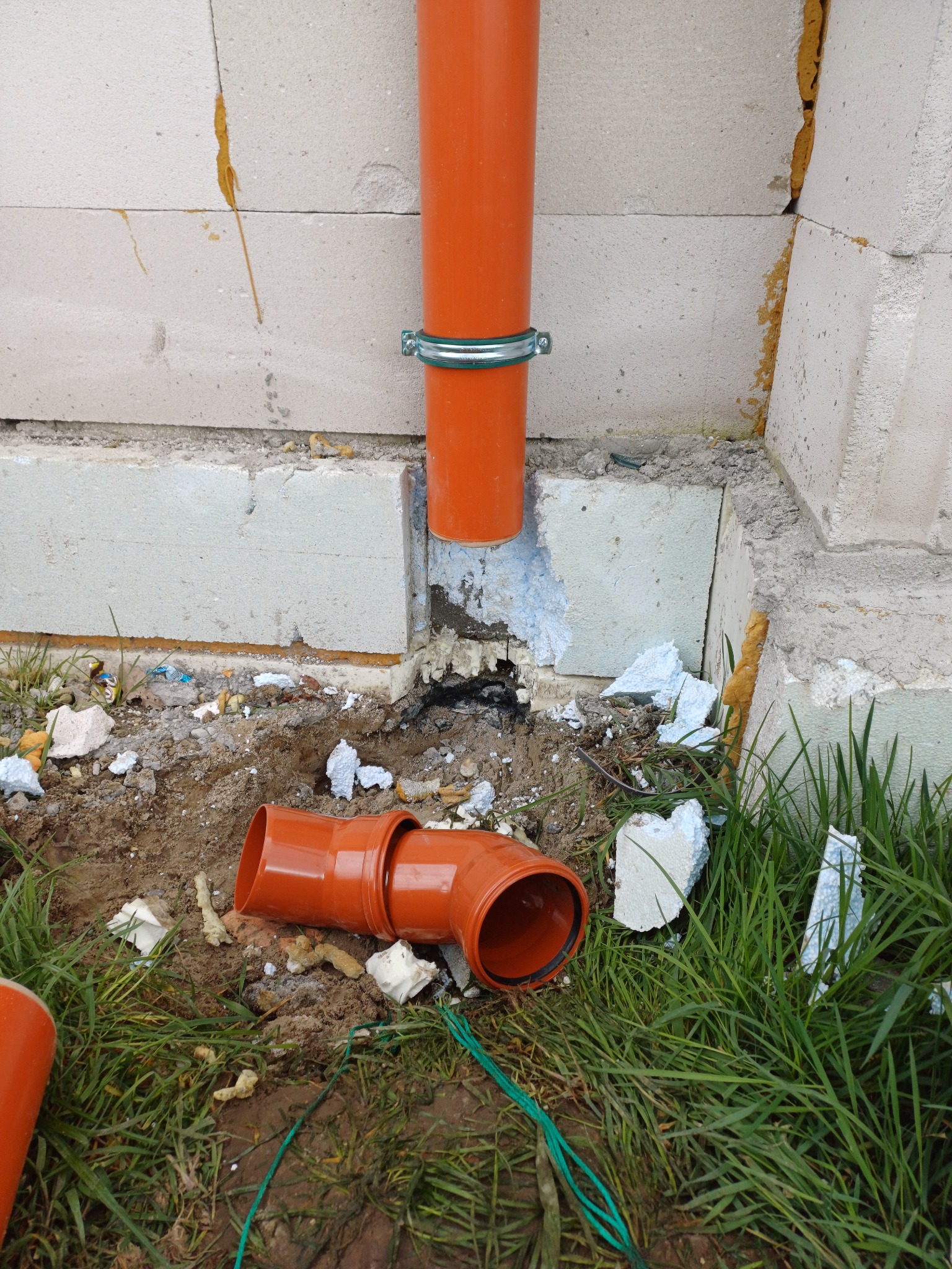 Pomarańczowe rury kanalizacyjne przy fundamencie z bloczków betonowych, widoczne fragmenty styropianu i pianki montażowej w ziemi.